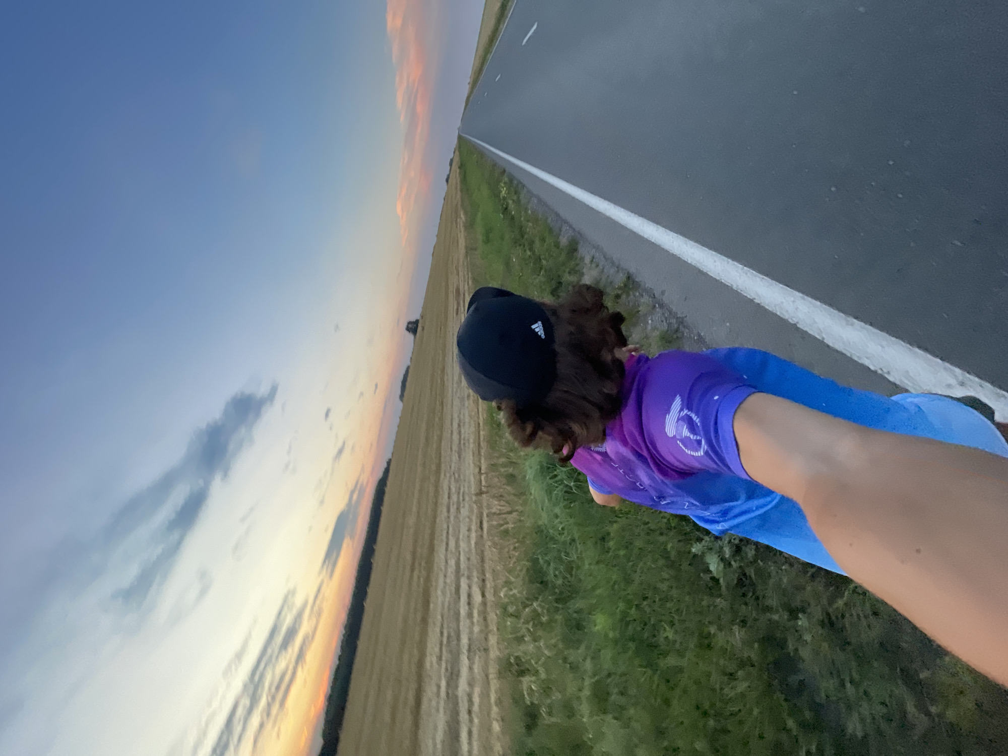 A first-person perspective image captures a person wearing a black Adidas baseball cap and a brightly colored, gradient shirt in shades of purple and blue, as they stand on the side of a rural asphalt road. The road stretches into the distance, marked by a white line. To the left of the road, a grassy verge is bordered by a harvested wheat field, with a line of trees visible in the distance. The sky above is a gradient of blue and pastel pinks and oranges, indicating either sunrise or sunset. The scene appears to be set in a rural area, possibly in the city of Ol'khovo, Russia, given the landscape and the agricultural field.
