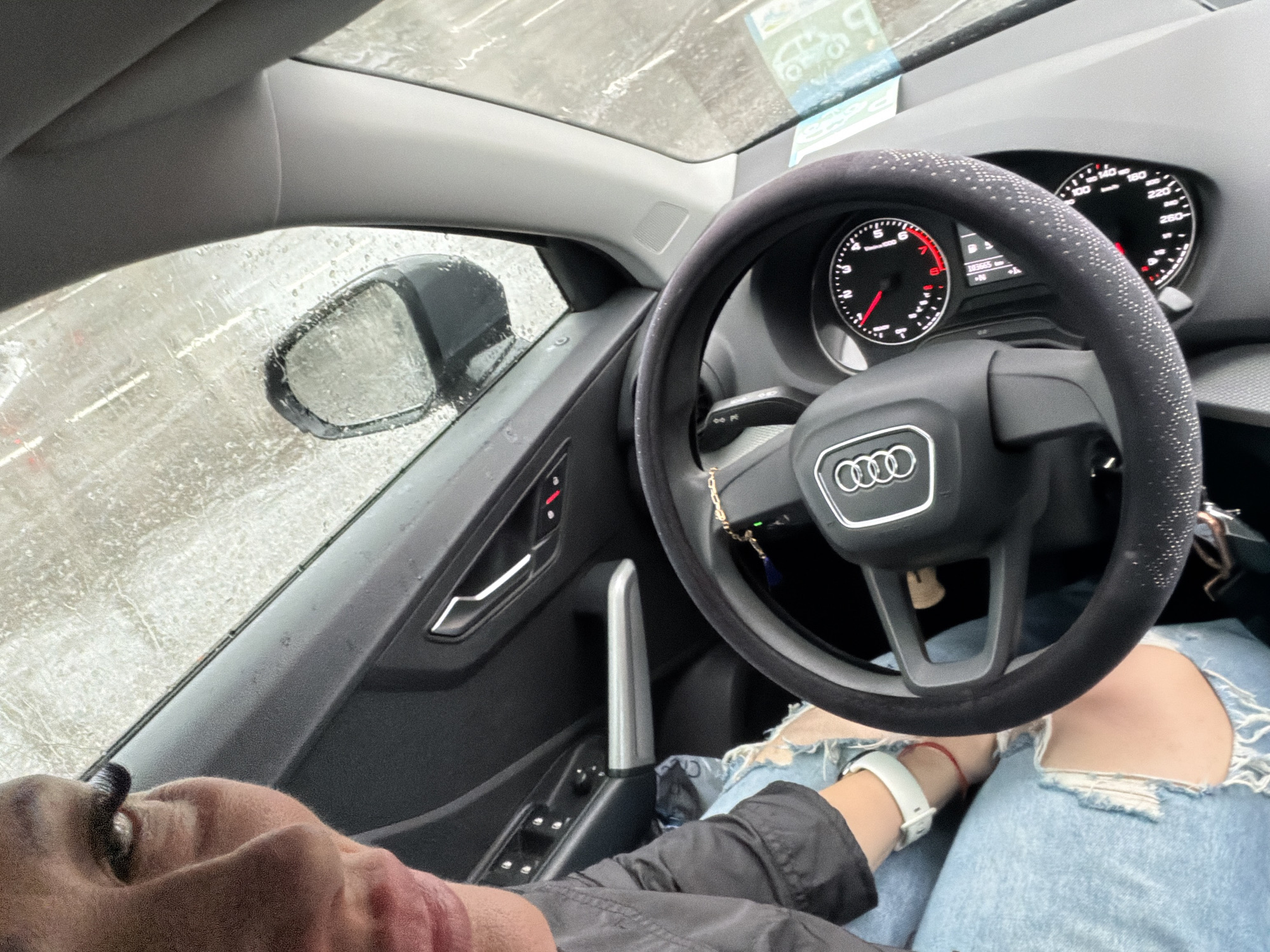 A low-angle indoor shot from the perspective of the driver of an Audi car, presumably in a city location, as suggested by the rainy or snowy street conditions visible through the car windows. The driver's face and part of their torso are visible in the lower left and center of the frame. They are wearing light blue ripped jeans, a white watch, and a dark-colored jacket. The car's interior is predominantly dark, and the steering wheel has a studded cover. The dashboard is visible, as is the side mirror reflecting the outside environment. The image appears to be taken in the daytime under overcast conditions.