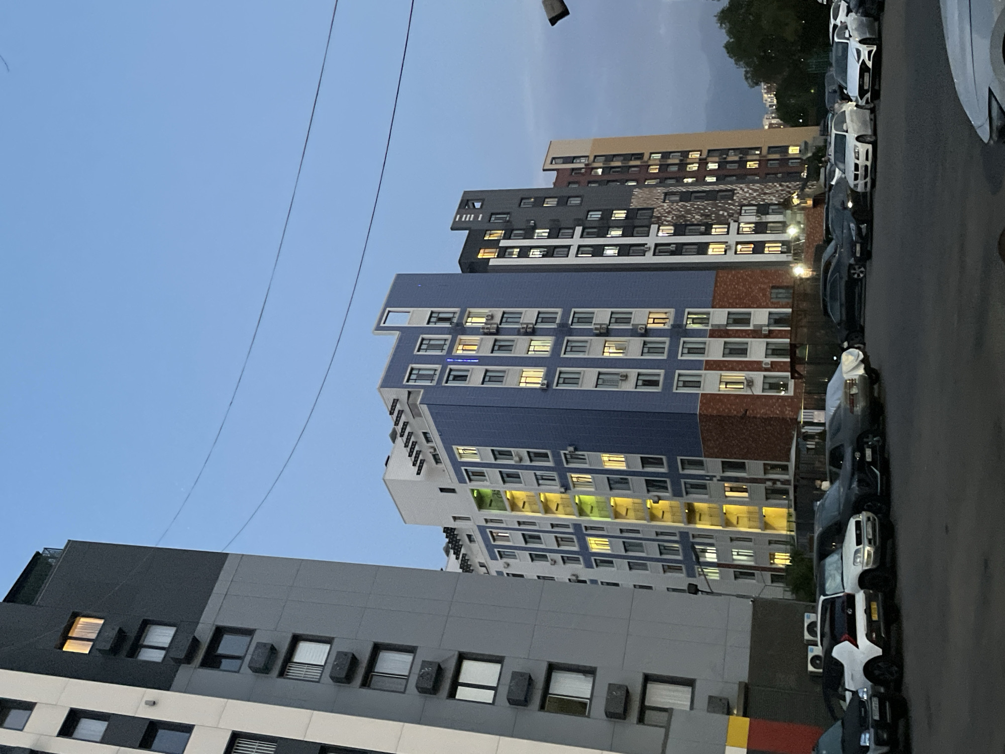 The image captures a street-level view of a residential complex in Almaty, Kazakhstan, during twilight. Multiple high-rise apartment buildings dominate the scene, characterized by their modern architectural designs featuring a mix of grey, white, blue, and beige facades. Several buildings have visible air conditioning units mounted on their exterior walls, and numerous windows are illuminated, suggesting occupancy.
In the foreground, a parking lot is filled with various cars, including white sedans and SUVs, a dark-colored SUV, and a black sedan. The vehicles are parked in rows. In the background, trees and other buildings can be seen, with the sky a pale blue, indicating the transition from day to night. Power lines are also visible stretching across the sky. The overall impression is one of a contemporary urban residential area.