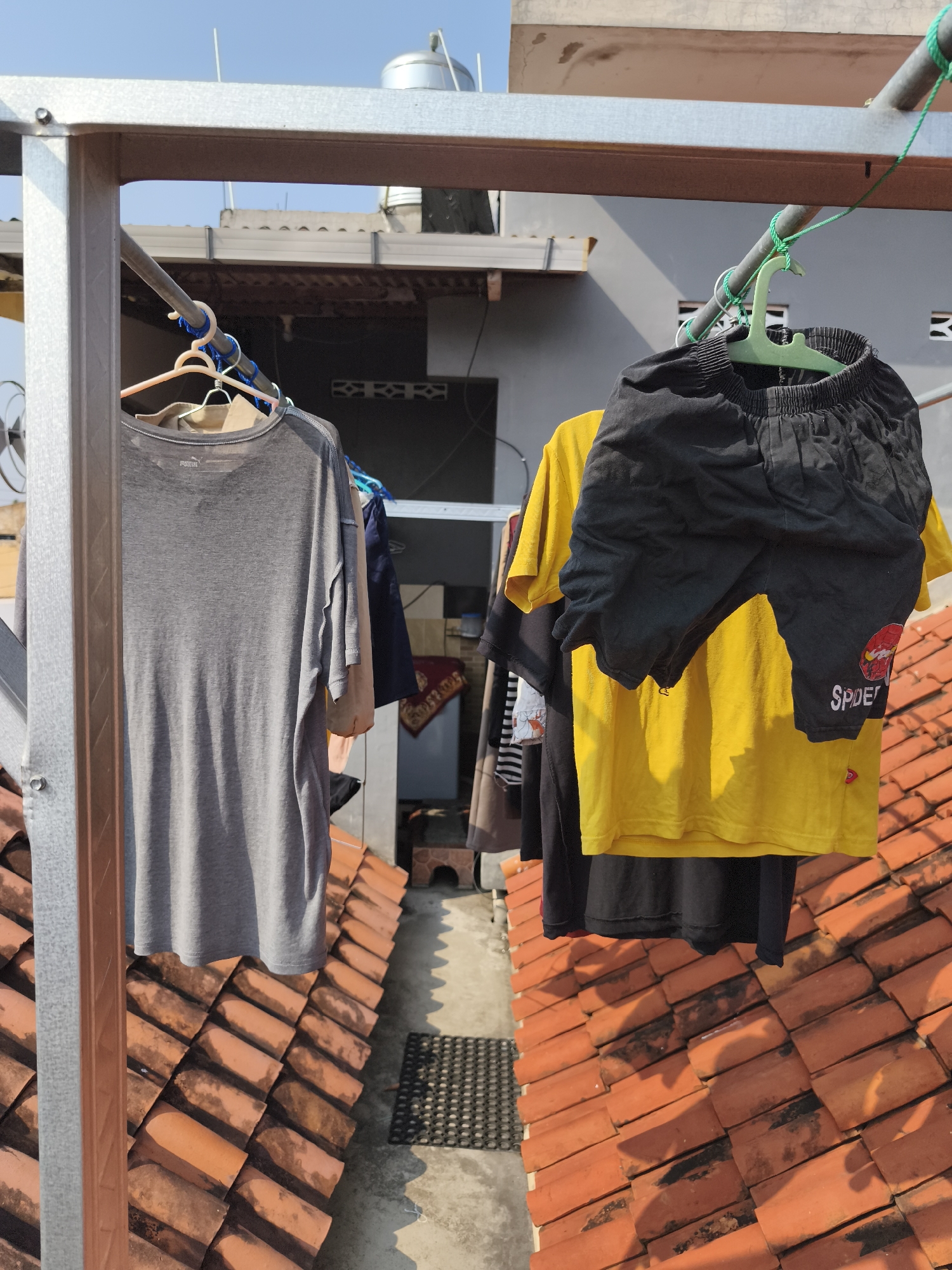A medium shot captures a clothesline outdoors, featuring several garments hung to dry. A grey t-shirt is on the left, a yellow t-shirt is in the middle, and black shorts are on the right. The clothes are hung on a metal frame above a tiled roof. In the background, there are buildings and a clear blue sky. This scene likely takes place in Yogyakarta, Indonesia, given the architectural style of the buildings and the prevalent use of tiled roofs. The lighting suggests it is daytime, likely a sunny day based on the shadows cast by the clothes.
