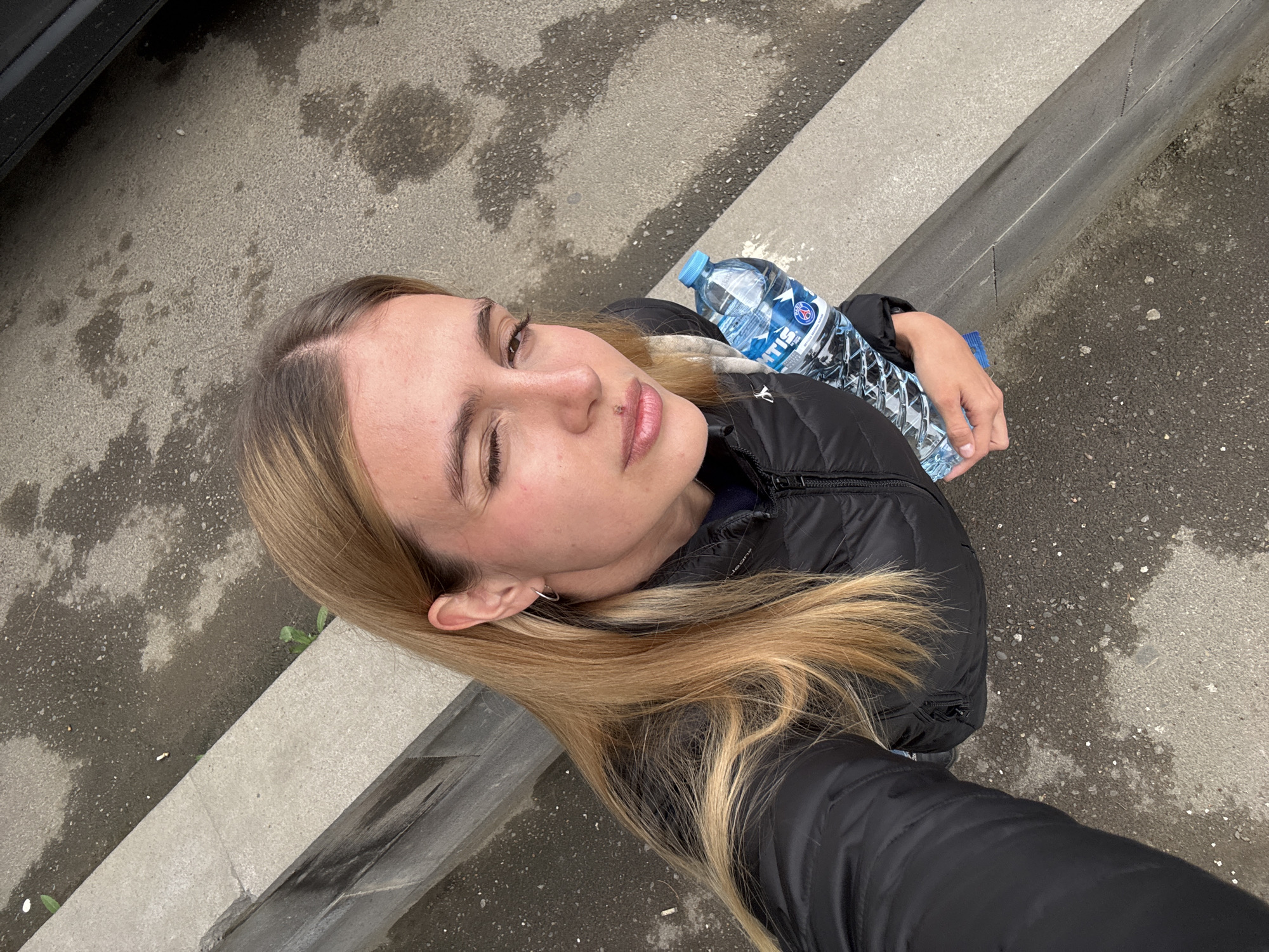 A woman in a black puffer jacket holds a bottle of water while standing on a city street. The street consists of a concrete sidewalk and asphalt with water stains. The location appears to be outdoors, with the woman positioned near the edge of the sidewalk next to the asphalt road. A car can be partially seen in the top left corner of the image. The woman's hair is blonde and shoulder-length.
