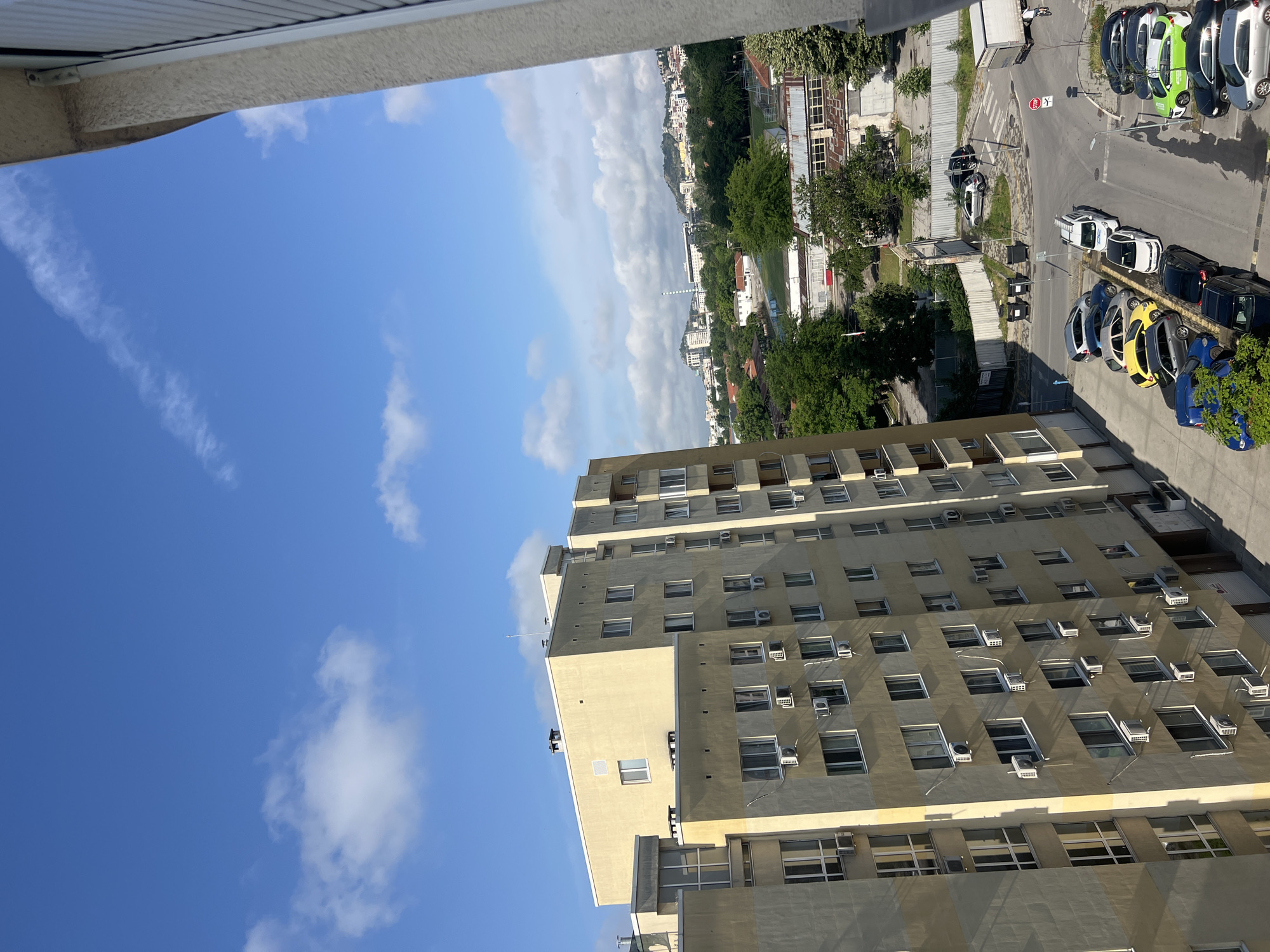 The image captures an elevated view of an urban landscape in Plovdiv, Bulgaria, on a clear, sunny day, likely during the morning or afternoon. The scene is dominated by a bright blue sky, speckled with scattered white clouds ranging from wispy trails to more substantial, fluffy formations.
In the foreground, on the right side of the frame, a tall, multi-story building with a light beige or yellow facade stands prominently. Its exterior is characterized by numerous rectangular windows, many of which have individual air conditioning units installed below them, reflecting the sun's brightness.
Below and to the left of this building, an active urban area unfolds. A parking lot or street is visible, populated by a variety of parked cars in diverse colors such as white, black, green, yellow, and blue. A white truck is also parked, and a lone figure on a bicycle or motorcycle can be seen near it, indicating a touch of human activity. A red 'STOP' sign is clearly visible near the roadway.
Further into the mid-ground, the urban expanse continues with a mix of other buildings, lush green trees, and some industrial structures, including a tall, slender chimney or tower. The background reveals more distant cityscapes, blending into the horizon where hills or mountains are faintly visible. The overall impression is a bustling city under a bright sky.