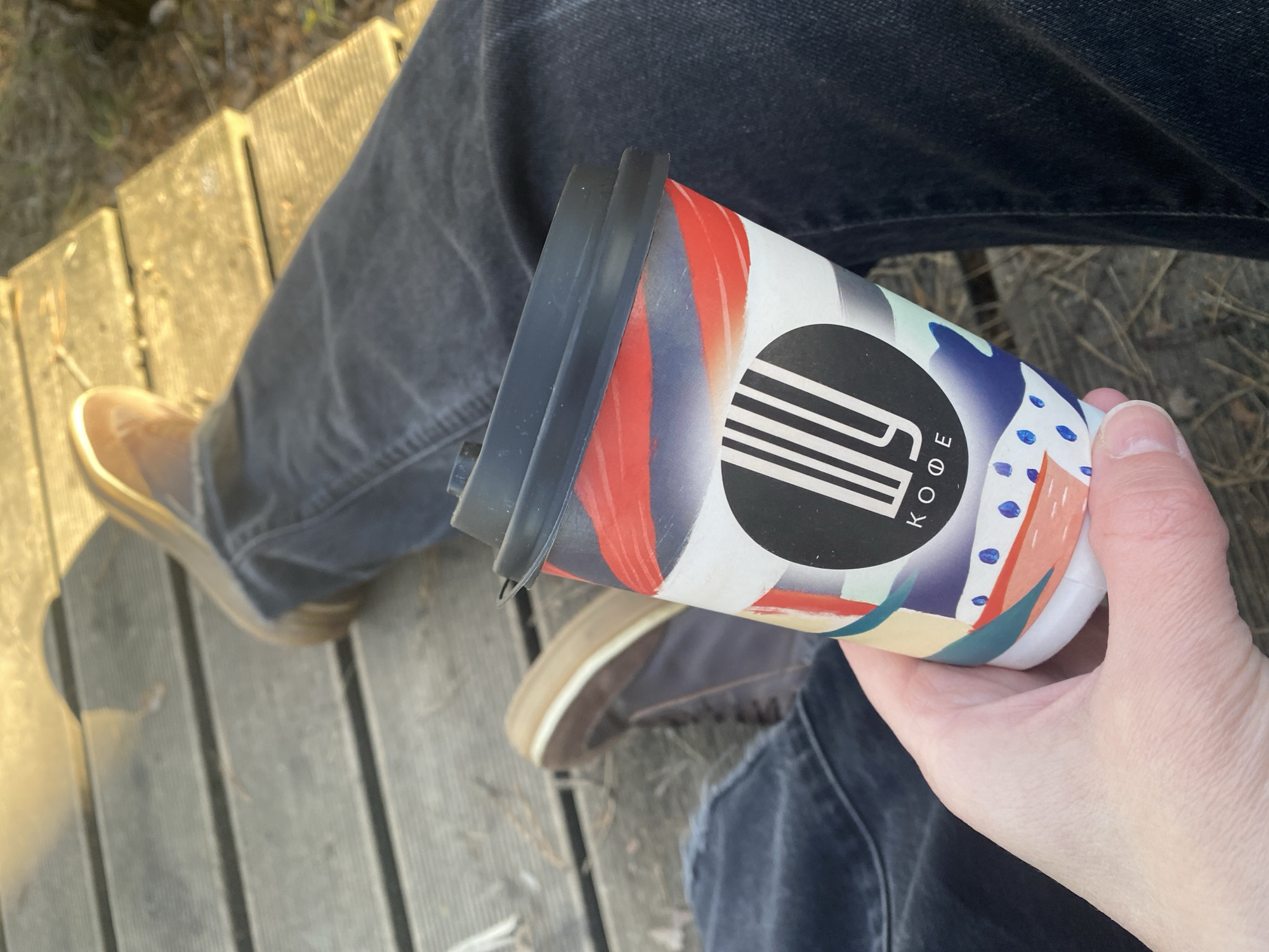 A person is sitting on a wooden deck, holding a paper coffee cup in their right hand. The cup has a black lid and is decorated with abstract patterns and the word "KODE" in Cyrillic. The person is wearing dark gray jeans and brown shoes. The deck is made of wooden planks and appears to be outdoors. I have no evidence in the image to determine the location in Chaikovsky, Russia.