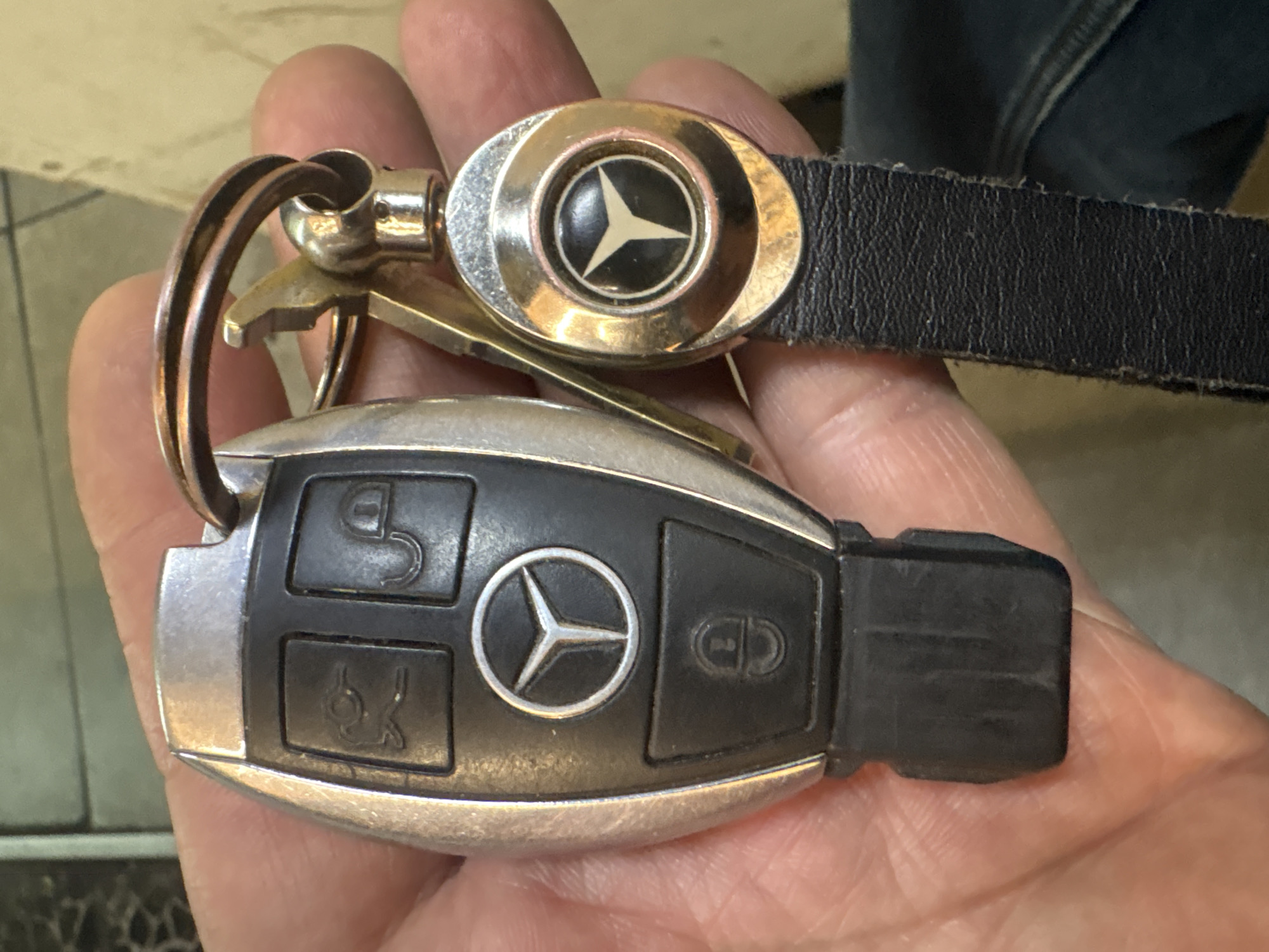 A close-up image shows a hand holding a Mercedes-Benz car key. The key is black and silver, with the Mercedes logo prominently displayed. Attached to the key ring are a metal bottle opener-style key and a Mercedes-Benz branded leather strap with a silver oval emblem. The background is out of focus, but jeans are visible, suggesting the person is wearing them. The location of Krasnoyarsk, Russia is not discernible from the image.