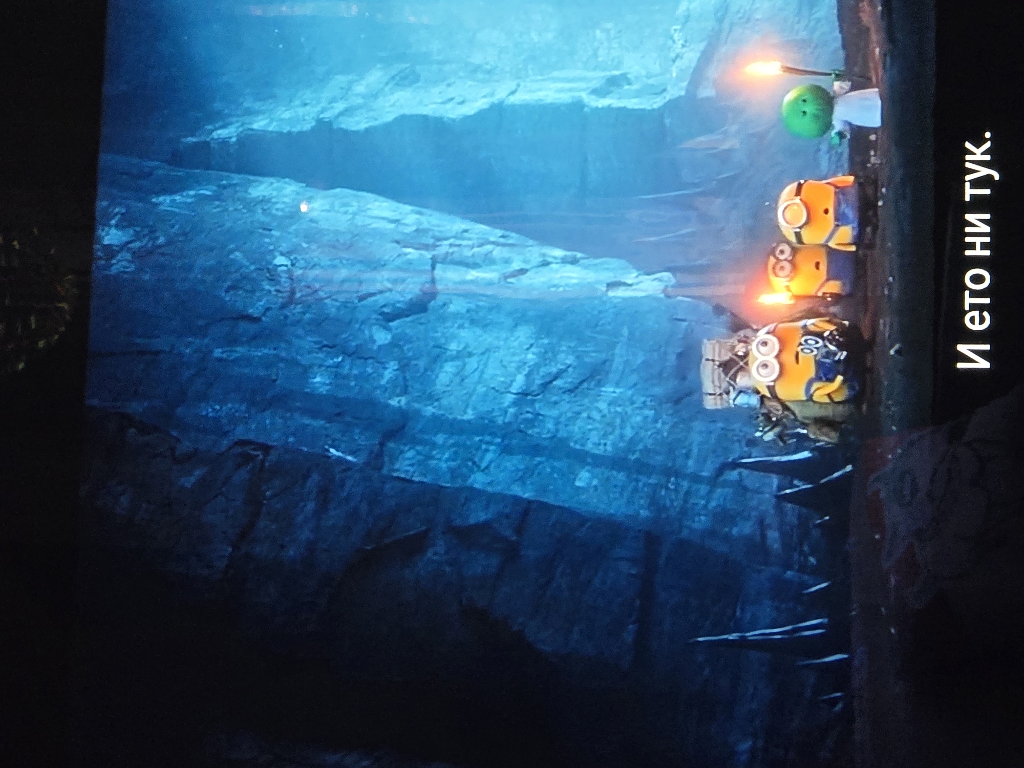 In a dark cave with jagged, blue-tinged rock formations, three Minions are illuminated by torches they are carrying. The Minions are small, yellow, cylindrical creatures wearing overalls and goggles. One Minion in the center appears to be looking at something off-camera with a surprised expression. To the right of the Minions, a small, green, spherical creature wearing a white robe is holding a torch. At the bottom of the image, white text in Cyrillic reads "И ето ни тук." which translates to "And here we are." The overall scene depicts the Minions and their companion exploring a cavern.