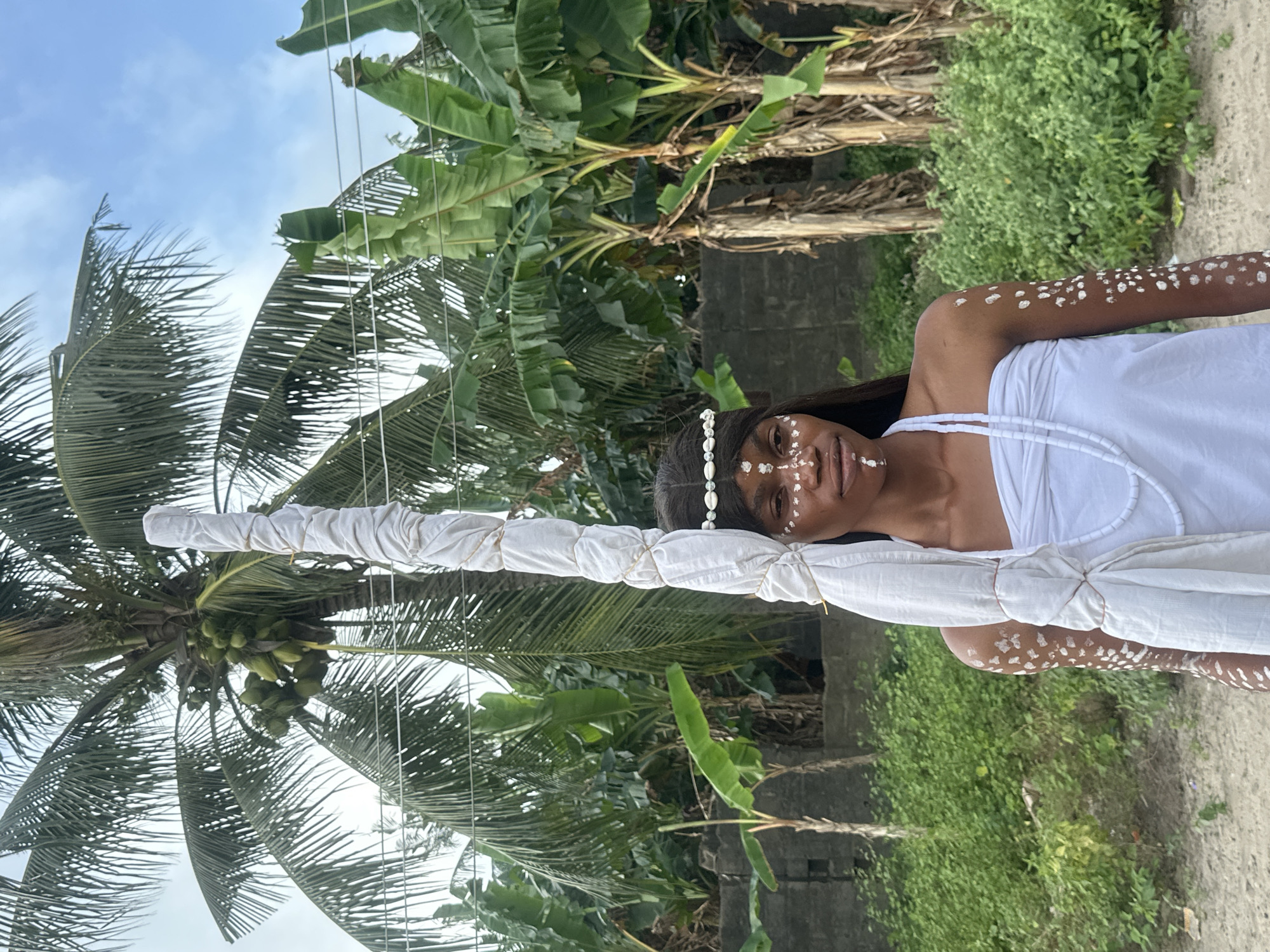 A young Black woman stands in a dirt area outdoors, posing for a photo in front of lush green vegetation and palm trees, with a cloudy sky overhead. She is adorned with white body paint on her face and arms, and wears a white beaded necklace and a headband made of shells. Her hair is dark and long. She is draped in white fabric, and a tall, white-wrapped object, possibly a staff or symbolic item, extends upwards next to her, secured with rope. The background includes banana trees, a low brick wall, and some overhead wires. The setting appears to be in or around the city of Badagry, Nigeria.