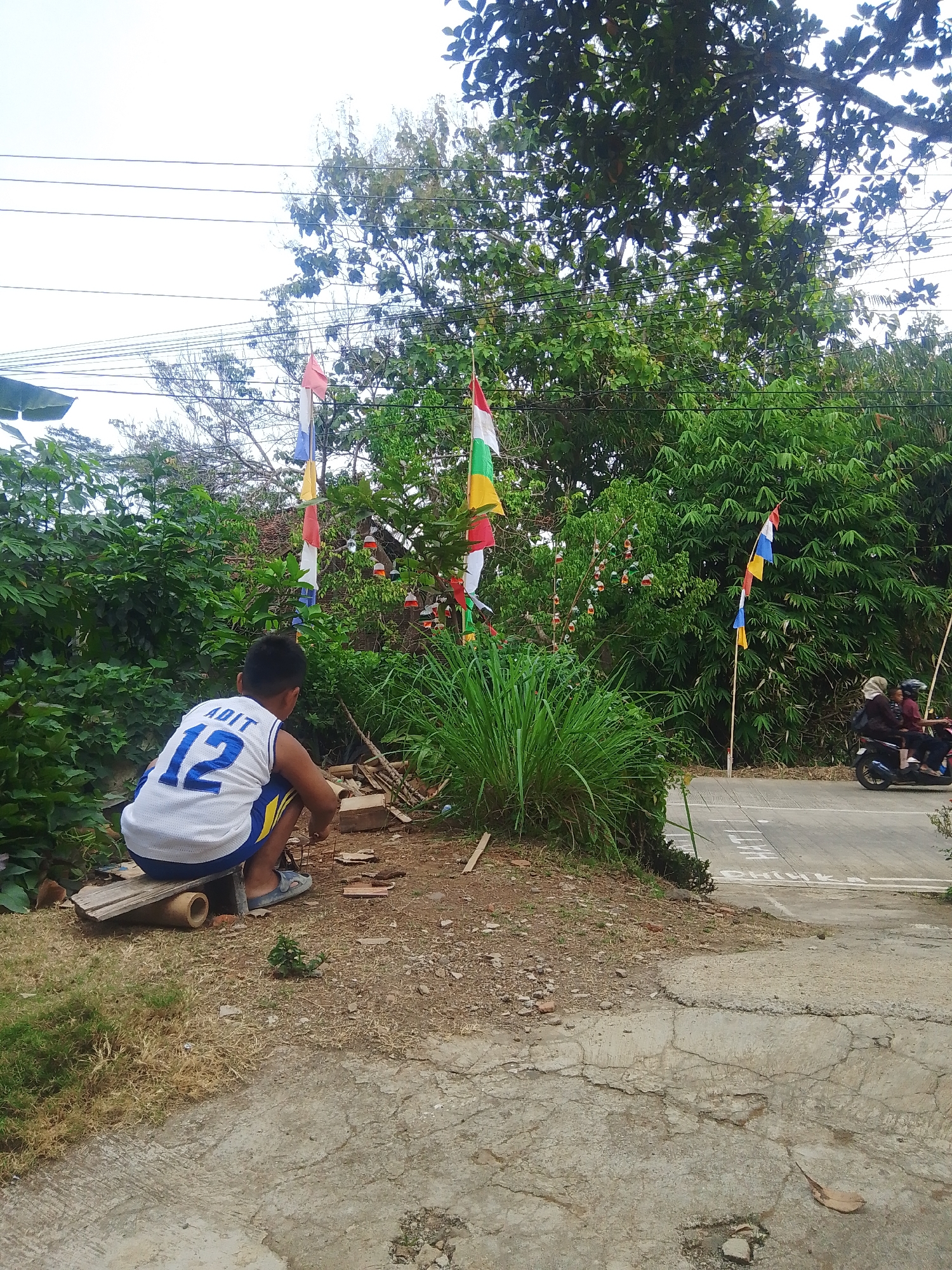 The image depicts an outdoor scene, likely in Bantar, Indonesia, characterized by lush greenery and clear skies. In the foreground, a young boy wearing a white basketball jersey with the number 12 and the name "ADIT" on the back is crouched down on a wooden plank. He appears to be engaged in some activity, possibly working with small objects. Surrounding the boy are scattered wooden debris and dry grass. Behind him, a variety of trees and bamboo plants form a dense backdrop. Several flags of various colors, including red, white, blue, and yellow, are visible, some attached to poles and others strung along wires, suggesting a festive or celebratory atmosphere. In the background, a paved road is visible, with a motorcycle carrying two individuals in the distance. The overall impression is a casual, everyday scene in a tropical environment.