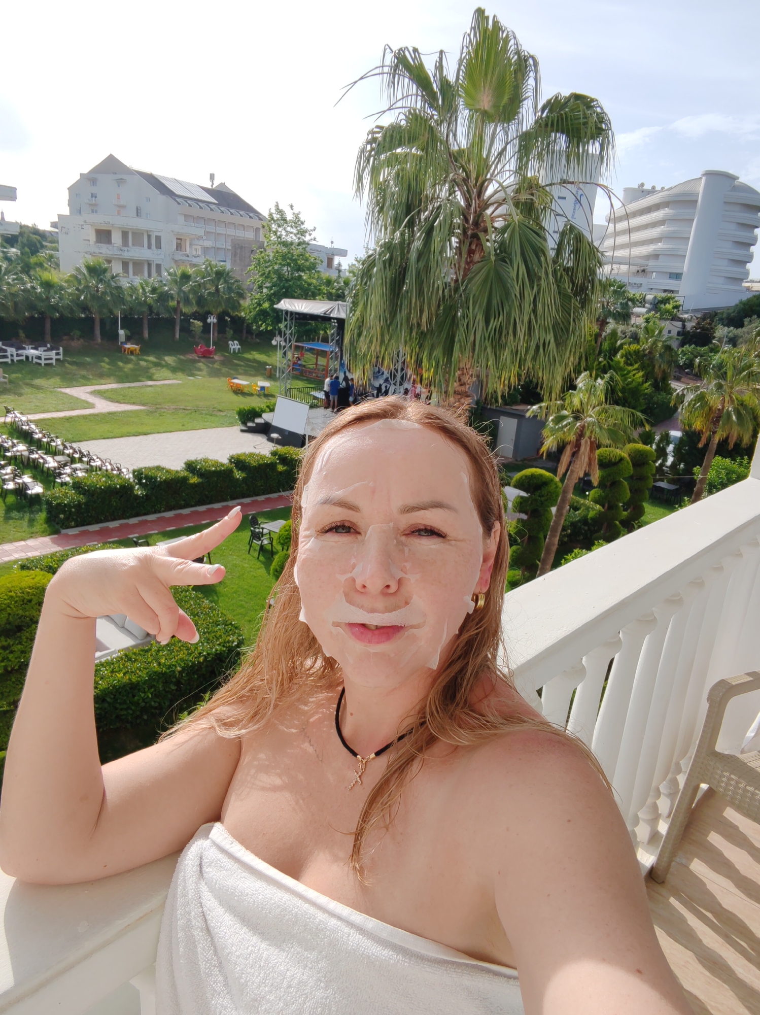 A woman with a sheet face mask poses for a selfie on a balcony. She wears a white towel, gold earrings, and a cross necklace. With her left hand, she makes a peace sign. The background shows a tropical resort landscape. A white building with architectural curvature stands on the right. A second, lower building on the left. A green lawn, palm trees, and other lush vegetation are visible. In the distance, lawn chairs are neatly aligned. A stage structure stands on the lawn. The scene suggests a sunny day at a hotel or resort in Alanya, Turkey.