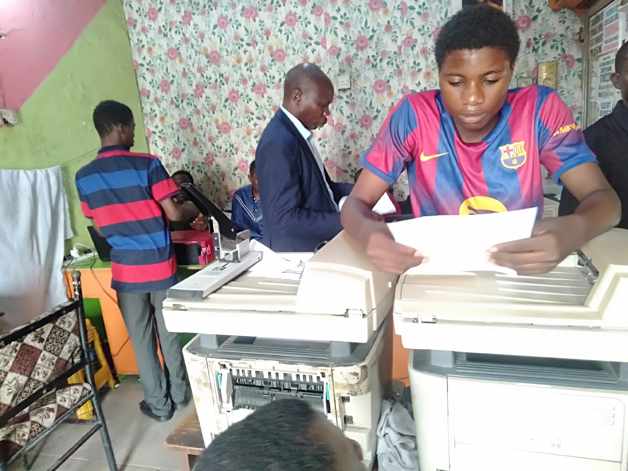 The image depicts a cluttered office space in Shangisha, Nigeria. A young man wearing a FC Barcelona jersey is standing in front of a white copy machine, holding a white sheet of paper. To his left, a man in a suit is also standing near a copier. Another man wearing a striped shirt is standing near a desk where a stapler is located. The room has a green and pink wall, and floral wallpaper. A patterned chair is visible.