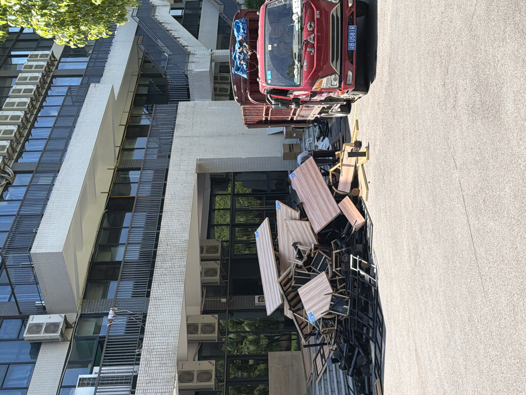 In front of a multi-story building with a tiled facade and numerous air conditioning units mounted on the exterior, a large pile of discarded furniture, including tables and chairs, is located on asphalt pavement. A red JAC truck is parked to the right of the debris. Two individuals appear to be in the process of loading the furniture into the truck. The building has multiple floors with balconies, large windows, and an external staircase on the right side. The sky is clear and the scene appears to be lit by natural daylight. The location is identified as Jiangjin, China.