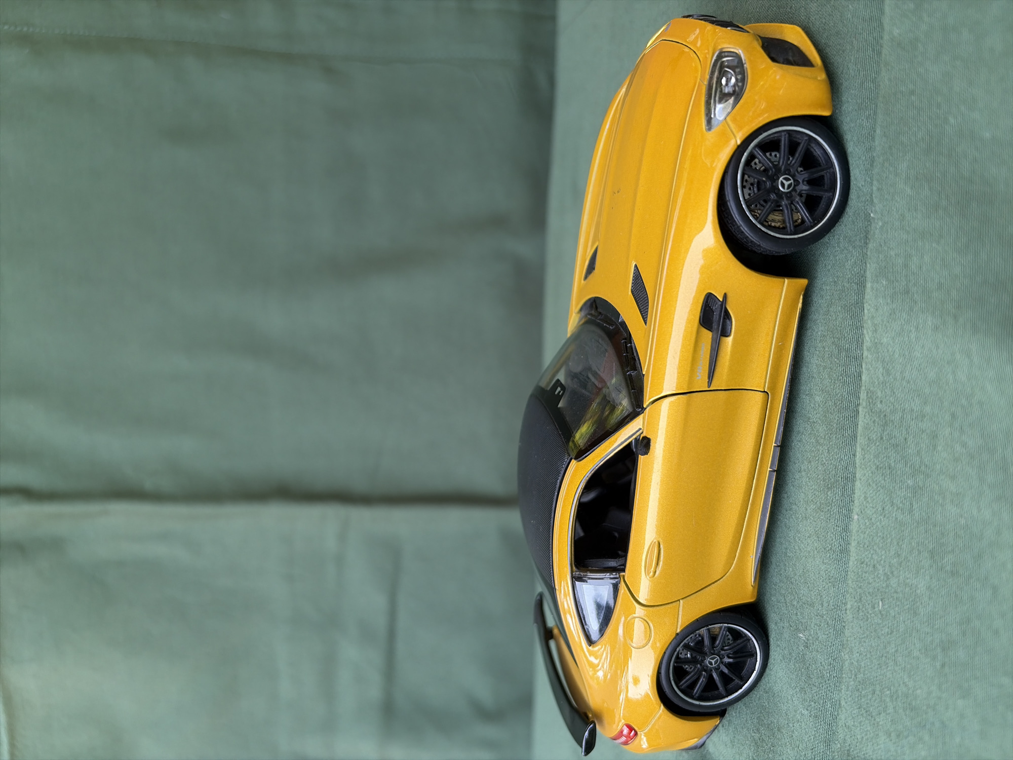 A die-cast model of a yellow Mercedes-AMG GT S is positioned on a dark green textured fabric surface. The car is viewed from its left side, angled slightly towards the front. It features a black roof, black wheels with silver accents, and a black rear spoiler. The background consists of a dark green fabric with visible vertical seams, suggesting it might be a draped cloth. The image does not contain any people, actions, or interactions, and there is no discernible urban environment or specific location within Oxentea, Moldova.