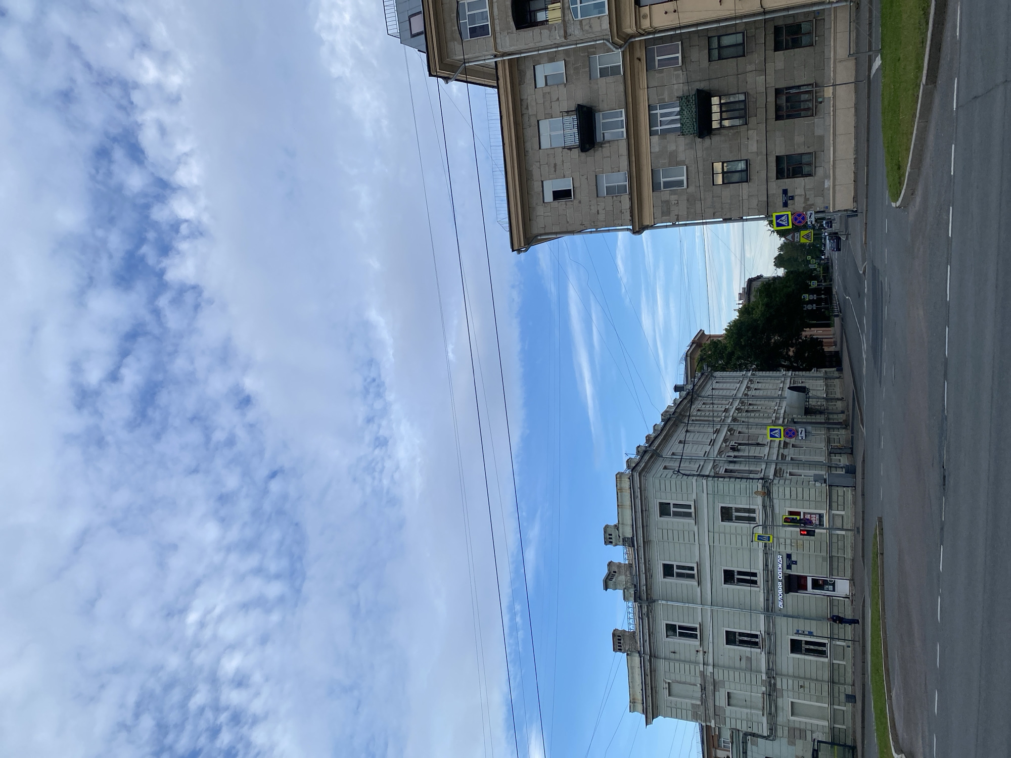 The image shows a street scene in Saint Petersburg, Russia, on a day with a partly cloudy sky. In the foreground is a paved road with white lane markings. On the left, a light blue, multi-story building with several windows and three chimneys on the roof dominates the scene. A sign above a doorway reads "ДЕЛОВАЯ ОДЕЖДА" (Business Clothing). A single person in dark clothing stands near the entrance. On the right, a gray, multi-story building with a more modern appearance stands. It has multiple windows, some with balconies, and a mansard roof. Traffic lights and pedestrian crossing signs are visible at the intersection. Further down the street, trees and more buildings line the road. Overhead, several power lines crisscross the sky.