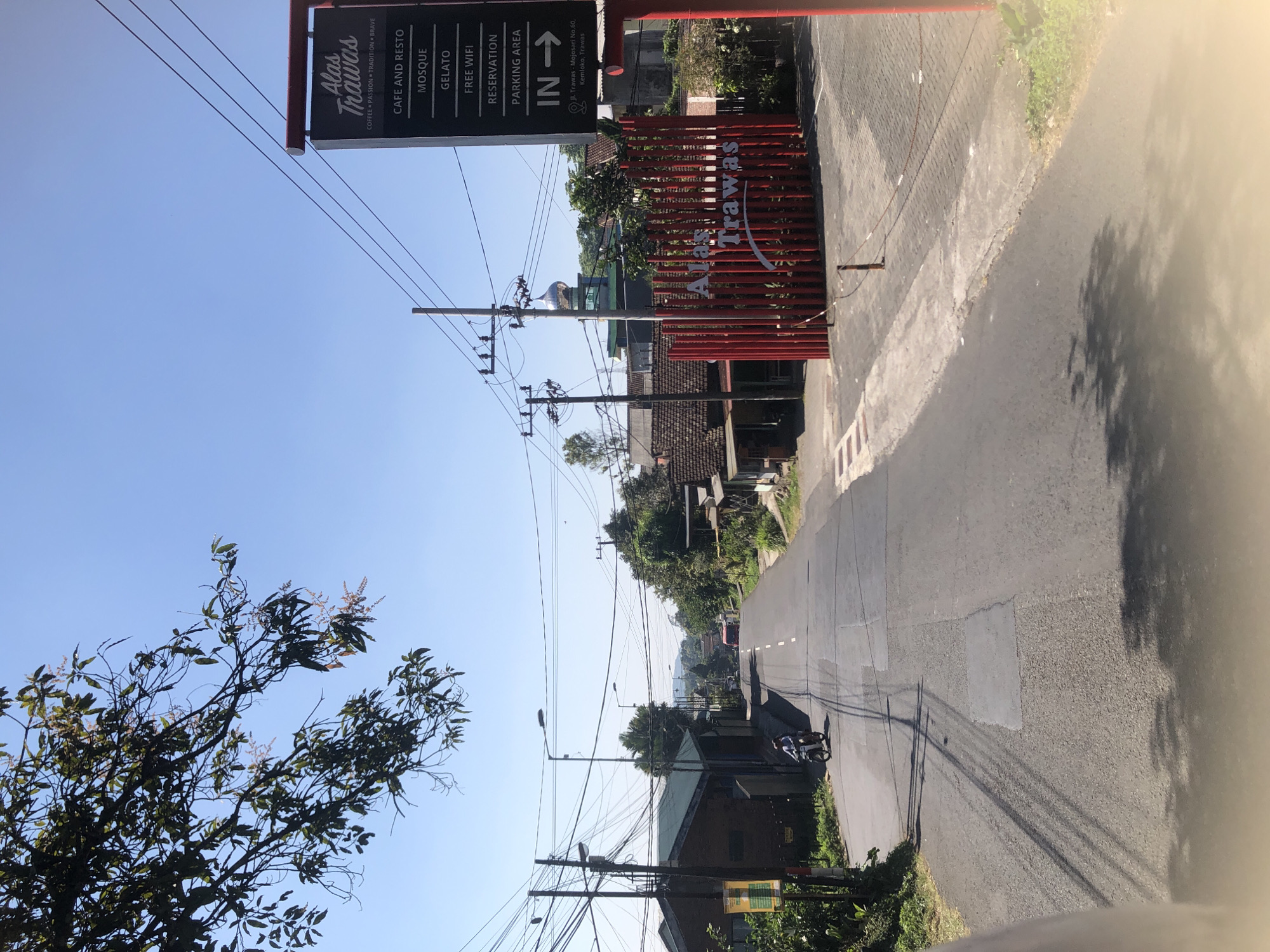 A street view shows a road leading into a village with a bright blue sky overhead. On the right side of the image, a red fence encloses a property with a sign that reads "Alas Trawas". The sign lists amenities such as "Cafe and Resto", "Mosque", "Gelato", "Free Wifi", "Reservation", and "Parking Area". Below this, an address is given: "Jl. Trawas - Mojosari No. 60, Kemloko, Trawas". A motorcycle with a rider is traveling down the road. In the background, a mosque with a silver dome is visible, along with houses and trees. Electrical wires crisscross the scene, and a tree branch extends from the top left corner. The road surface appears to be asphalt with some patches and tire marks. The scene depicts a sunny day in Trawas, Indonesia.