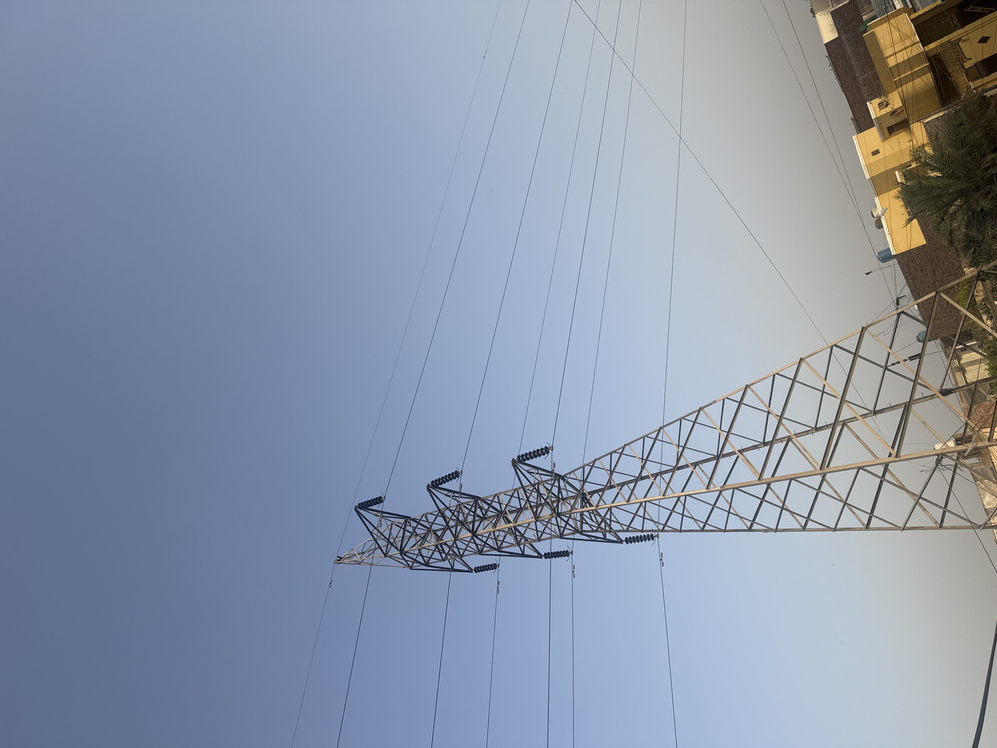 An upward-angled view captures a large, multi-tiered metal lattice electricity pylon prominently positioned against a clear blue sky. The sky transitions from a deeper blue at the top left to a lighter, slightly hazy blue-grey towards the lower right horizon, with no visible clouds. Numerous dark power lines extend horizontally across the sky, connected to the pylon via multiple sets of dark, disc-shaped insulators. Several small, dark specks, possibly birds, are faintly visible in the sky.
The pylon, constructed from interconnected metal beams, exhibits a weathered metallic grey or light brown appearance and tapers upwards. It occupies the central and left portions of the image, rising from below the frame to nearly the top edge.
In the lower right quadrant, a section of an urban environment is visible. This includes a bright yellow residential building with a flat roof and small dark openings, partially obscured by a dark green tree or large bush with palm-like fronds. Adjacent and behind the yellow structure are buildings constructed from reddish-brown brick. A light blue water tank is discernable on one of the rooftops. Additional thin, dark wires crisscross above the buildings. The image exhibits a slight rightward tilt in its composition. No human figures or discernible animal life are present.