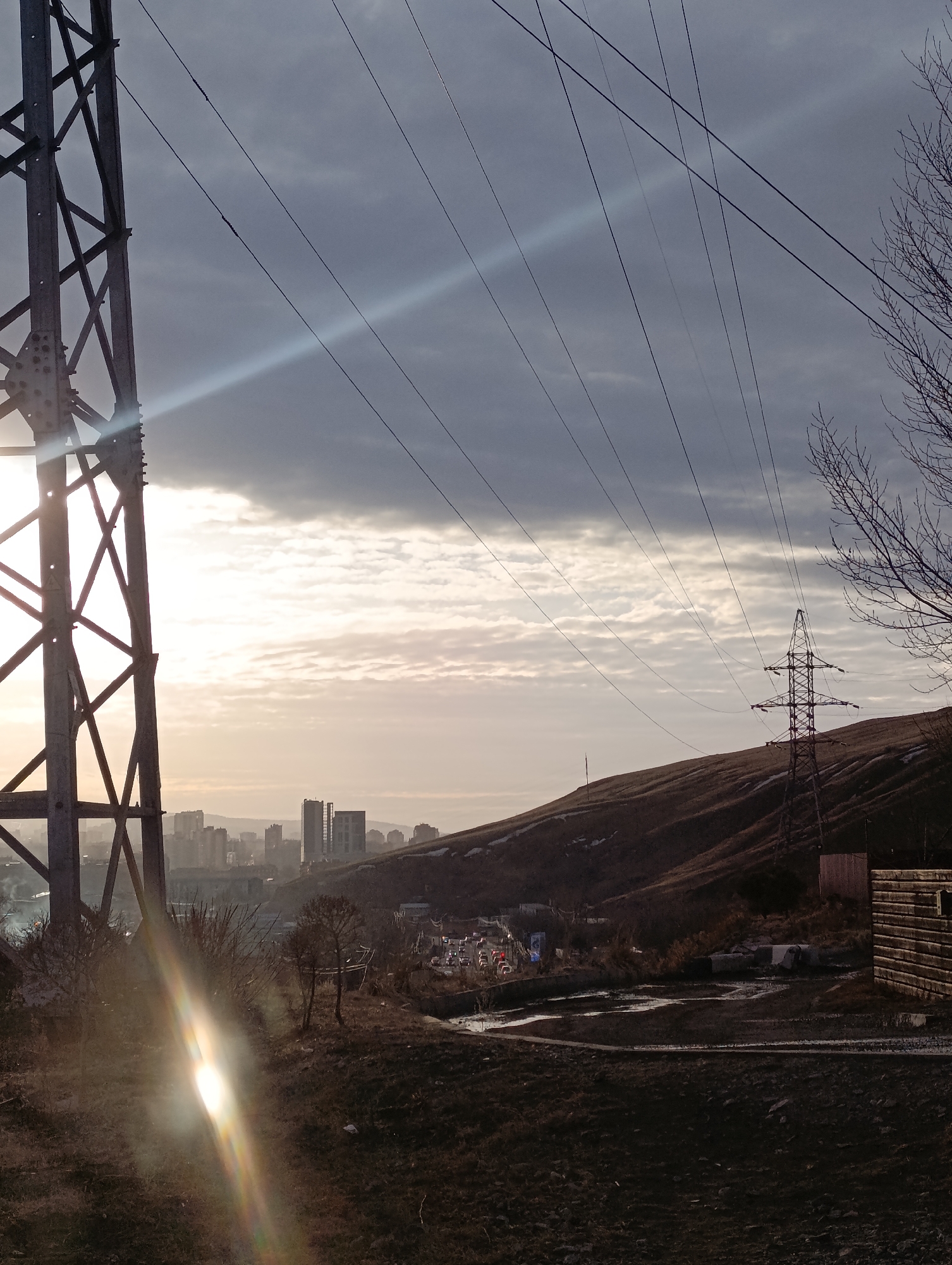 The image presents an elevated panoramic view within the city of Krasnoyarsk, Russia, captured during either dawn or dusk. In the immediate foreground, a large, grey metallic lattice transmission tower (pylon) stands on the left, partially silhouetted against a low, bright sun that causes a significant lens flare extending diagonally upward and to the right. Numerous parallel power lines originate from this pylon, stretching across the sky.
The ground in the central and right foreground is unkempt, composed of dark, uneven earth, dry brown vegetation, and visible puddles or wet patches. A rectangular wooden structure, appearing weathered, is situated on the far right.
In the midground, a hazy urban skyline of Krasnoyarsk is discernible, featuring multiple multi-story buildings and structures. Below this skyline and extending to the right, a road or highway is visible, populated with vehicles indicated by their illuminated headlamps and tail lamps. To the right, a large, barren hill with sparse dry grass and small patches of snow rises, dominating the midground and background. A second, smaller metal lattice transmission tower stands on the slope of this hill, integrated into the power line network.
The sky exhibits partial cloud cover, transitioning from bright, sun-illuminated areas near the horizon to darker, denser cloud formations at the top of the frame. No individuals are directly visible.