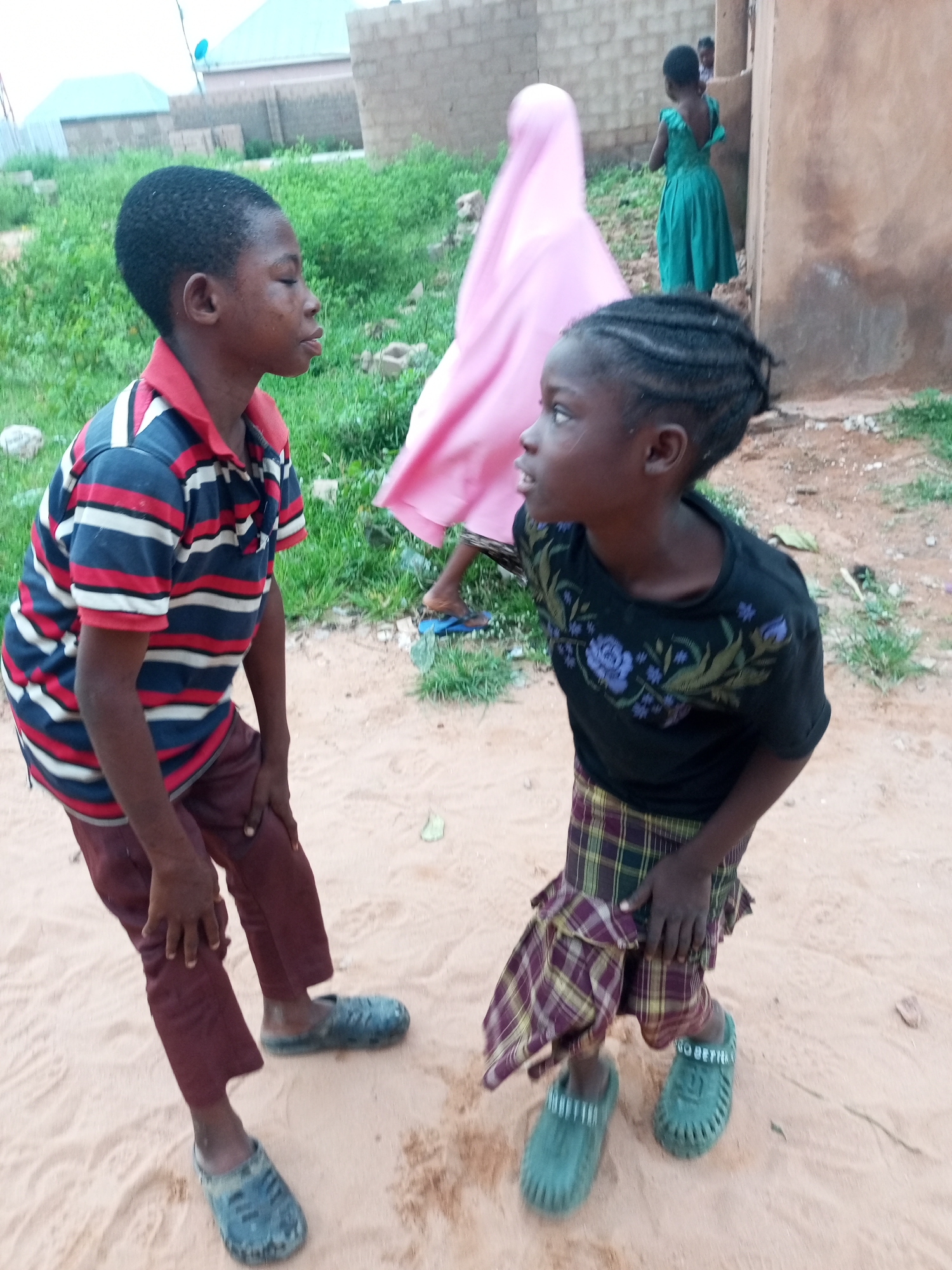 In Tempure, Nigeria, two young children stand on a sandy ground. The child on the left, a boy, wears a striped polo shirt in red, white, and blue, and maroon pants. He has his right hand on his knee. The child on the right, a girl, wears a black t-shirt with floral embroidery and a plaid skirt. She has her right hand resting on her left leg. Behind them, another girl in a pink hijab is partially visible, and further back, a girl in a green dress stands near a building. The environment appears to be an outdoor area with sparse vegetation and brick structures.