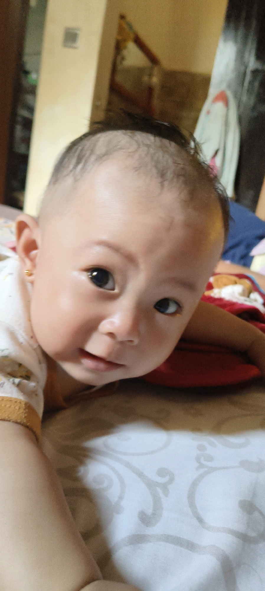 A close-up, shallow depth-of-field, eye-level shot focuses on the face of a baby lying down in Waisai, Indonesia. The baby's head is angled towards the viewer, with its body extending out of frame to the left. The baby has dark eyes, a small nose, and slightly parted lips. A small gold earring is visible in its left ear. Its hair is sparse and dark, with a shaved or very short cut on the sides of its head. The baby is wearing a white shirt with a yellow trim and a subtle pattern. The background is blurry, showing a bed with patterned sheets and a red pillow. The overall lighting is soft, with hints of natural light coming from the left.