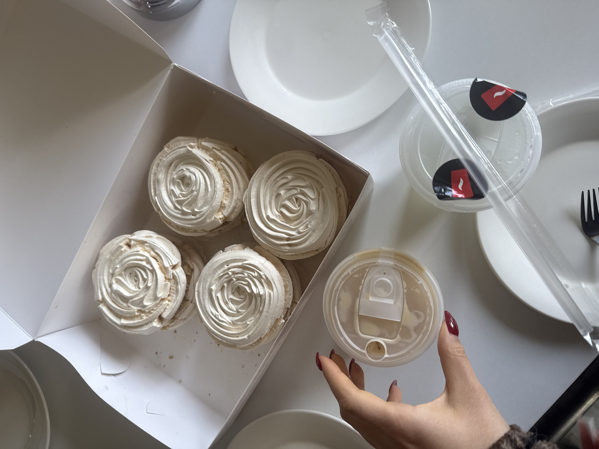 The image is a high-angle shot of a table setting featuring a box of meringues and beverages. A white cardboard box contains five meringues arranged in two rows. To the right of the box are several white plates, a clear plastic cup with two black and red labels, a clear plastic straw still in its wrapper, and a black plastic fork. In the foreground, a person's hand with dark red nail polish is holding a clear plastic cup. The overall scene is clean and well-lit, suggesting an indoor setting. There are no discernible details in the image indicating a specific location in Yerevan, Armenia.