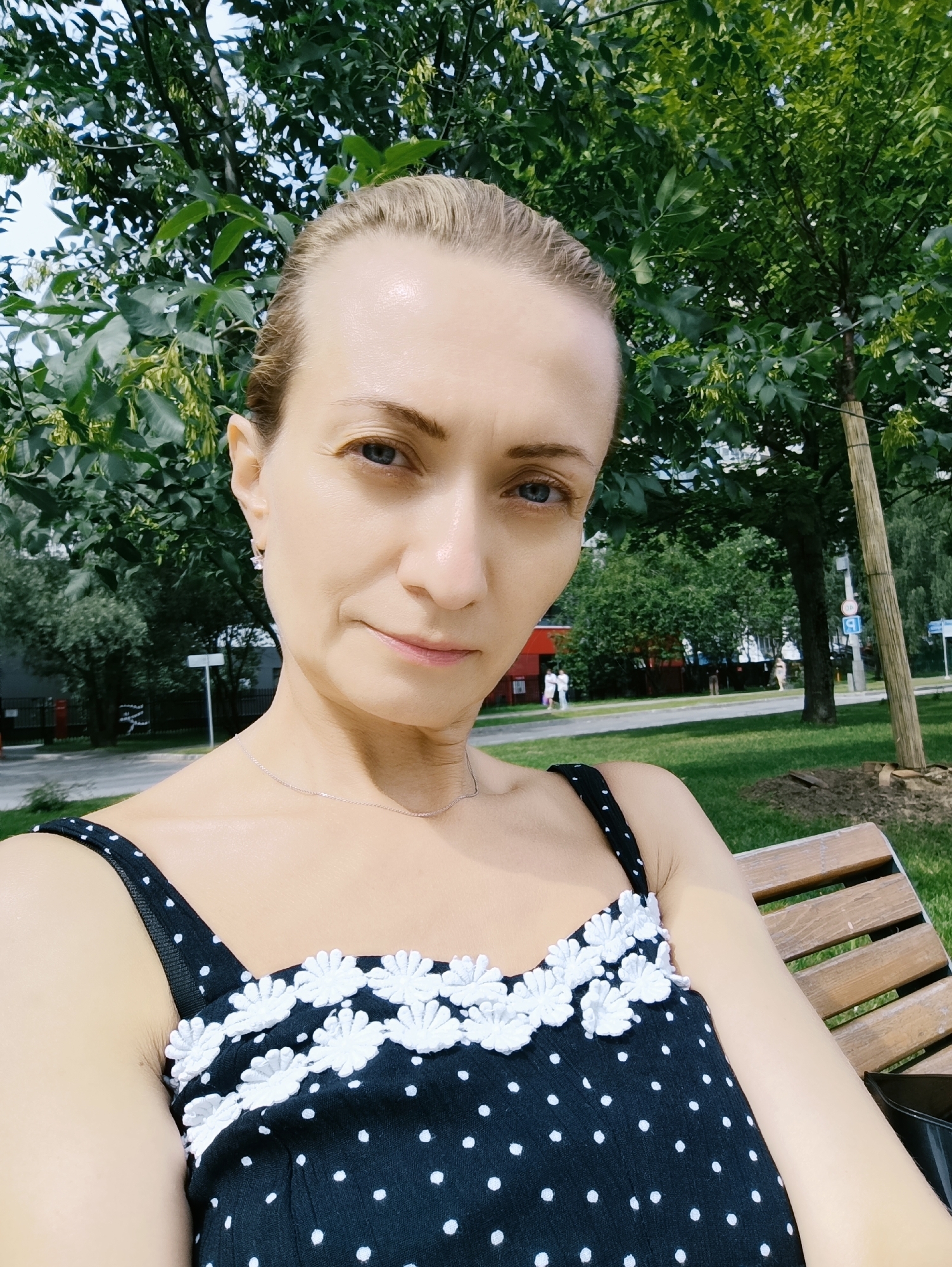 A woman is taking a selfie while sitting on a park bench. She is wearing a black polka dot dress with white flower appliques on the bodice. She has blonde hair pulled back and is looking directly at the camera. Behind her, there are trees, grass, and a street with buildings and some people walking in the distance. The scene appears to be in a park or public space within a city, potentially Moscow, Russia, given the context.