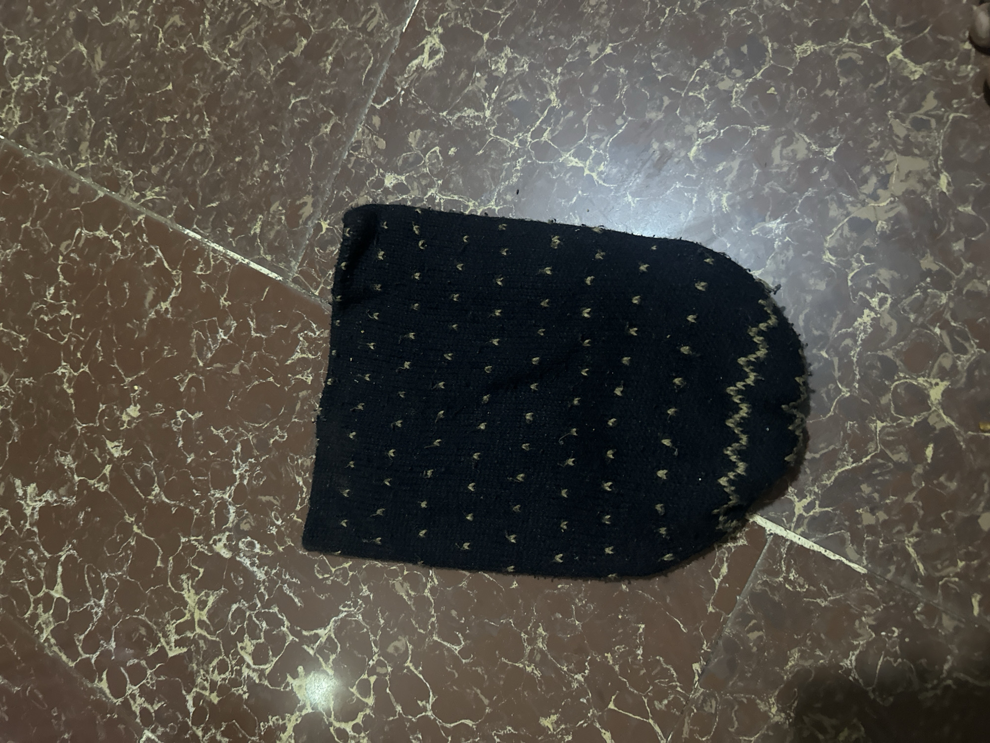 A dark knitted hat with gold speckles is lying on a marble tiled floor. The hat is oriented with its opening facing upwards and slightly to the left. The floor tiles are a reddish-brown color with lighter veins running through them.