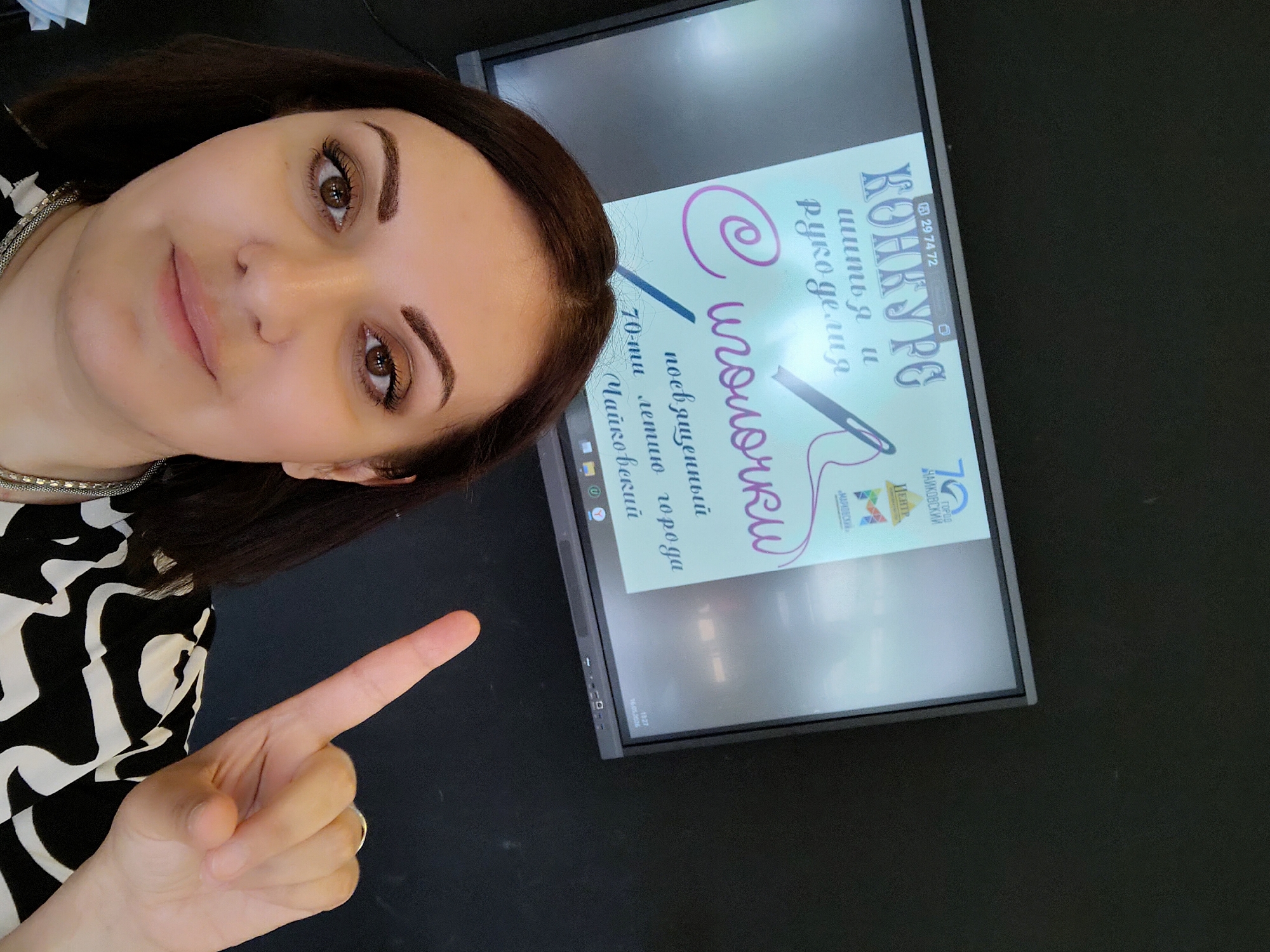 A woman poses for a selfie, pointing her finger towards a computer monitor displaying a poster. The poster advertises a "Needlework" competition dedicated to the 70th anniversary of the city of Chaikovskii. The text is in Russian. A logo featuring the number "70" and the words "City of Chaikovskii" in Russian is visible on the poster. The environment appears to be indoors.
