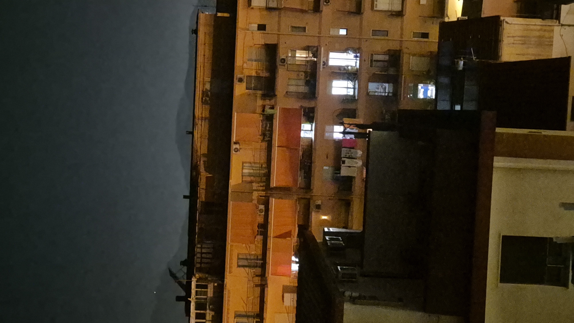 An exterior, medium shot captures a nighttime view of apartment buildings in an urban setting. The sky above is dark and overcast, with faint outlines of distant hills or mountains visible on the horizon. The buildings are multi-story structures with numerous windows, many of which are illuminated, suggesting occupancy. Several balconies are visible, some with awnings or closed shutters. Clothes hang on a line on one of the balconies, adding a touch of domesticity to the scene. The overall impression is one of a quiet, residential night in a city.