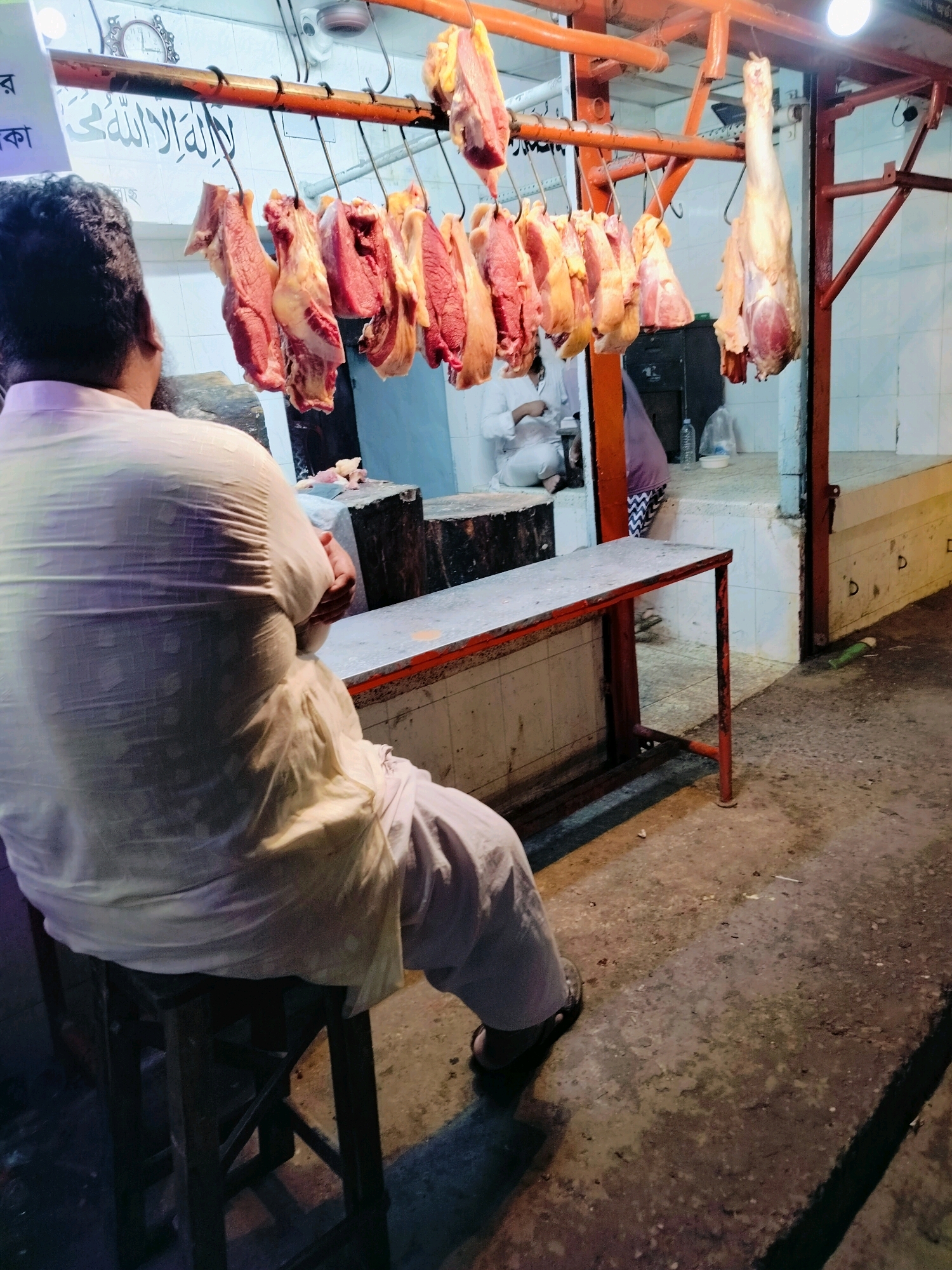 The image depicts an outdoor butcher shop located in Dhaka, Bangladesh. In the foreground, a man wearing a light-colored, loose-fitting shirt and pants is seated on a dark stool, facing away from the camera. He has dark hair and a beard. Suspended from an orange metal rack above, numerous cuts of raw meat hang by hooks. One large piece of meat, possibly a leg or hindquarter, is prominently displayed at the right end of the rack. Behind the meat, the shop's interior is visible, with tiled walls and a white counter. A person in a white garment is partially visible in the background, seated. The ground surface appears to be concrete or pavement, with some debris. A red-framed table with a metallic top is positioned in front of the counter. The lighting suggests it might be late afternoon or early evening.