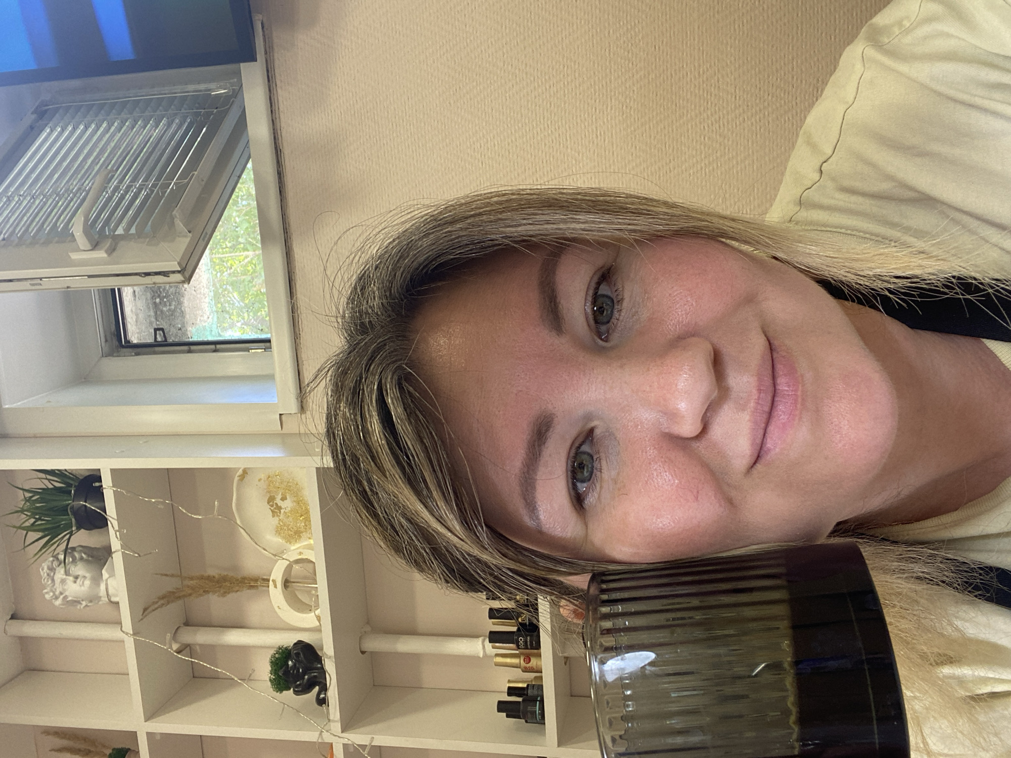 A medium shot captures a woman holding a dark glass cup in her left hand, which is positioned near her face. The woman has fair skin, light brown hair, and is looking towards the camera. Her lips are slightly parted, and she has visible blush on her cheeks. She is wearing a light yellow t-shirt with a black strap visible around her neck.
In the background, a white shelf unit is partially visible, filled with various decorative items. These include a white bust, a black pot with a green plant, a dried plant in a white vase, and a decorative round object with gold flakes. Further down the shelf, several small bottles, likely nail polish, are arranged. To the right of the shelves, a window is open with horizontal blinds partially closed. Outside the window, greenery is visible. The wall behind the woman appears to be a textured beige color. The scene suggests an indoor environment, possibly a home or a beauty salon, given the presence of nail polish bottles.
The provided context of "Chaikovsky, Russia" does not offer any visual cues within the image itself that would confirm or deny this specific location.
