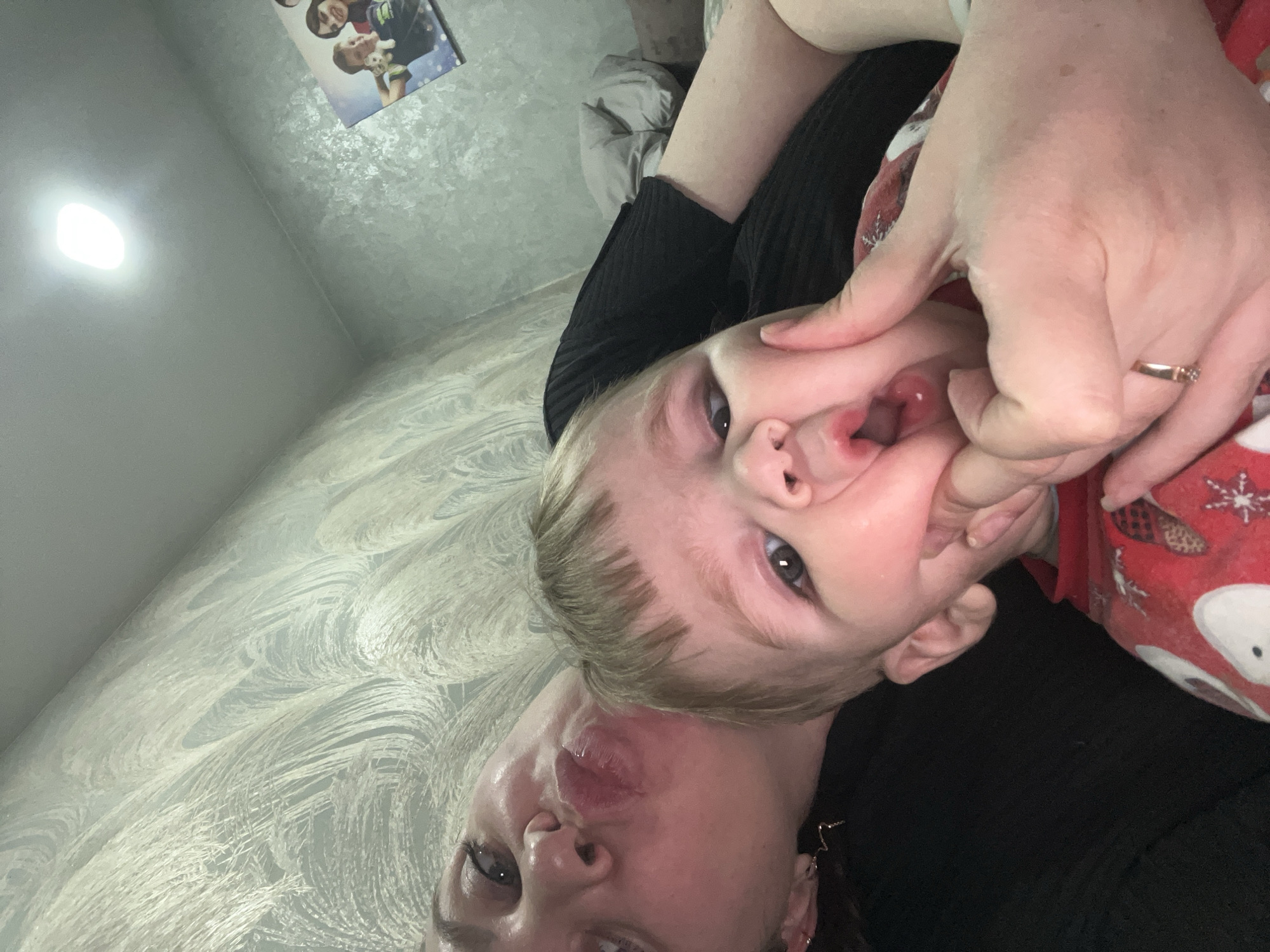 A selfie shows a woman and a young child, with the woman at the bottom and the child above her. The woman is pulling a funny face, puckering her lips. The child's face is being distorted by a hand that is holding his cheek. The background is a patterned wall. A photo is hanging on the wall. The location is unknown and there is no mention of Russia in the image.