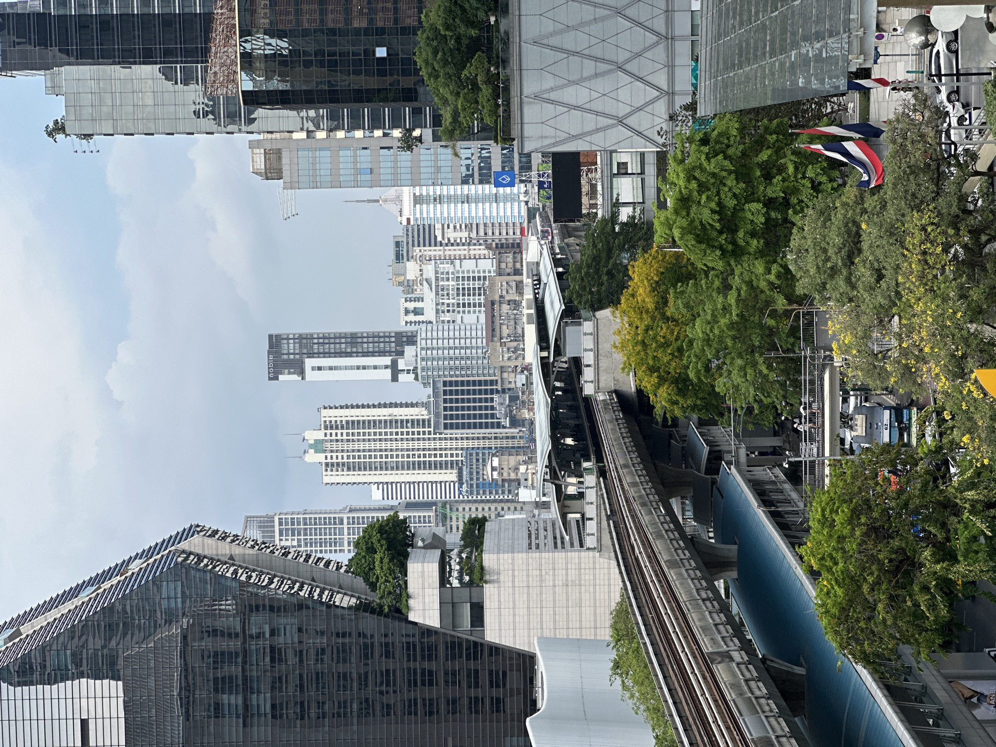 The image captures an elevated view of Bangkok, Thailand, showcasing a blend of modern architecture and natural elements. In the foreground, a section of the city's elevated train tracks runs parallel to a blue-roofed structure, with lush greenery surrounding the tracks and the buildings. Various high-rise buildings, characterized by their glass facades, dominate the skyline. The Thai flag waves amidst the trees, indicating the location. Street level traffic is visible, although obscured by foliage. The sky is overcast, casting a soft light across the urban landscape.