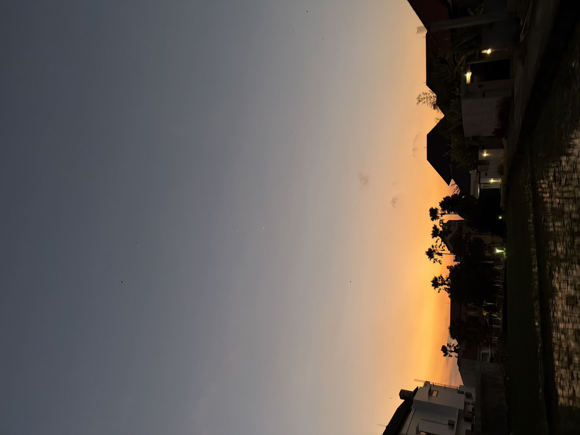 This image captures a sunset scene in North Kuta, Indonesia. The sky displays a clear gradient, transitioning from dark grey-blue at the top to a bright orange-yellow hue at the horizon, indicating dusk or post-sunset conditions. Multiple small, dark specks, consistent with birds or insects, are scattered across the upper and mid-portions of the sky. Two distinct bright yellow points of light are visible in the upper-right sky quadrant.
The foreground consists of a dark, reflective body of water, likely a flooded rice paddy, mirroring the sky's colors and the silhouettes of adjacent elements. In the midground, a distinct line of silhouetted structures and tropical vegetation is observable against the vibrant sky. On the left, a multi-story white building with a dark roof, external air conditioning units, and an exposed ladder or scaffolding is partially visible. Further right, numerous dark silhouettes of palm trees and other dense foliage are interspersed with several residential buildings. These buildings feature dark, sloping roofs and some exhibit localized illumination, indicating operational interior or exterior lighting. No human figures or discernible human actions are present within the frame.
