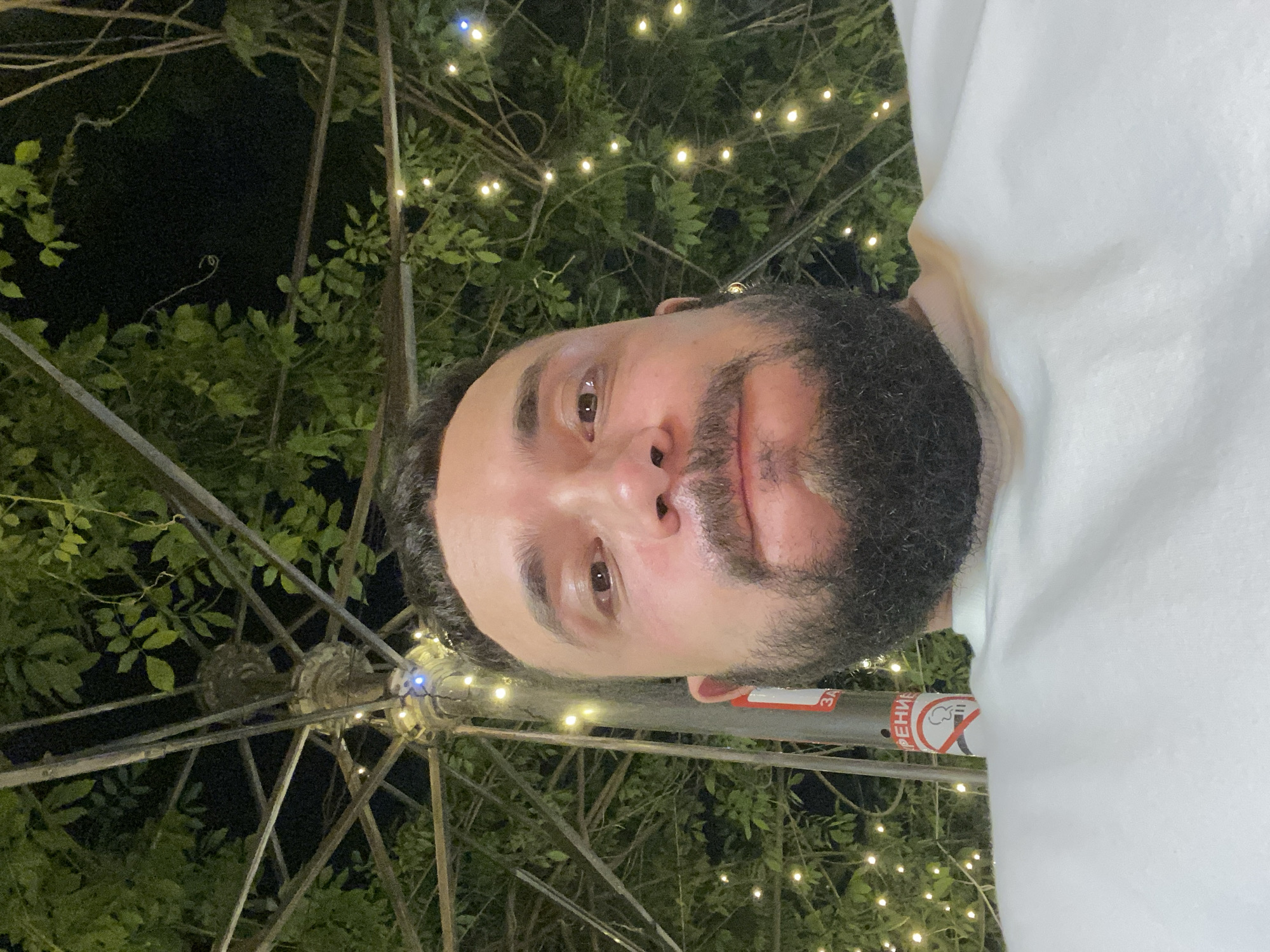A man with a beard takes a selfie at night in Makhachkala, Russia, under an umbrella-like structure adorned with string lights and foliage. He is wearing a white shirt and is positioned in the foreground, with a pole to his left featuring two stickers, one of which is a no-smoking sign. The background is dark with blurred green leaves and yellow lights.
