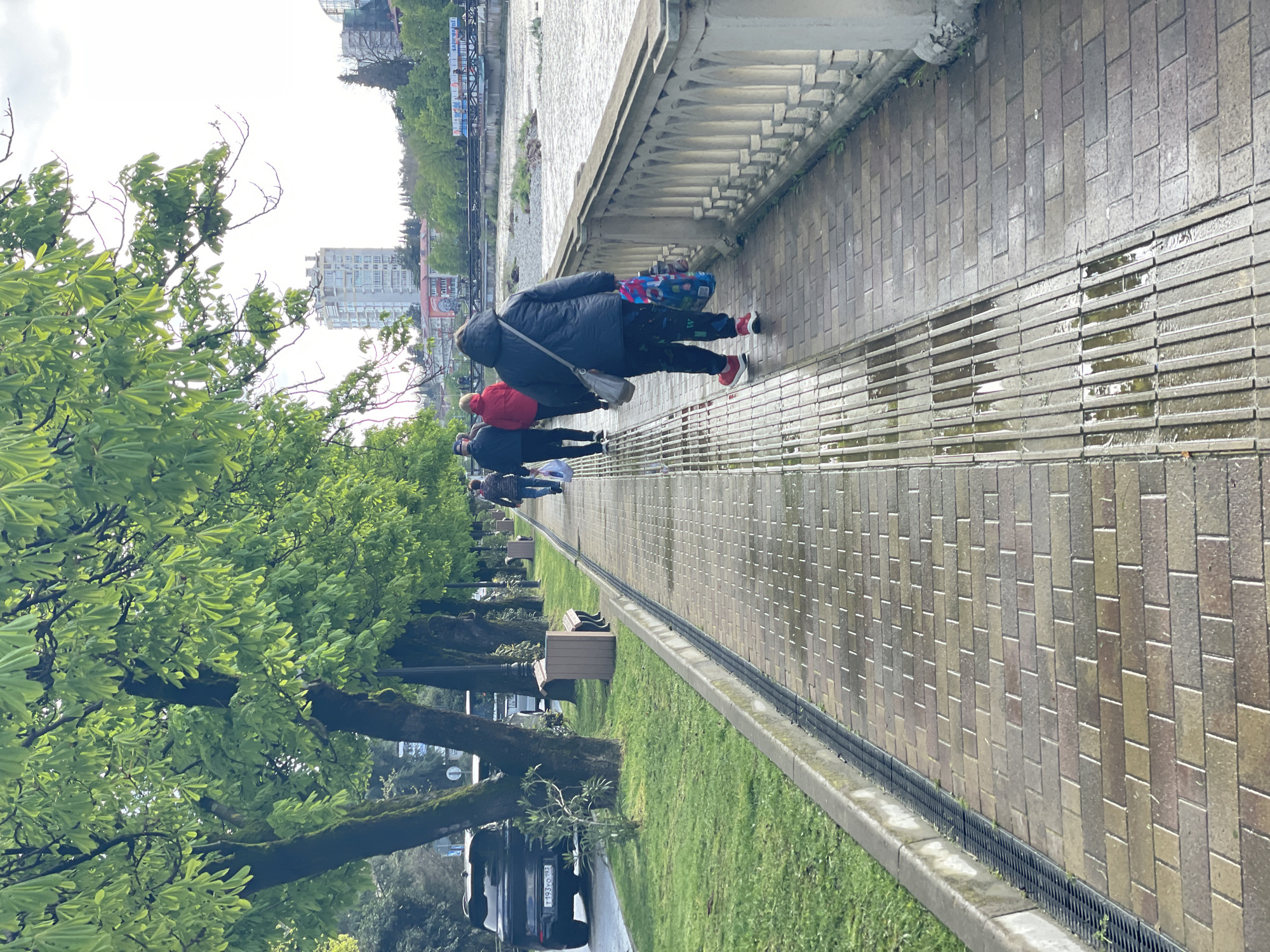 The image captures a street scene in Sochi, Russia, specifically on a walkway along the shoreline. Several people are walking along the brick-paved walkway, which appears wet from recent rain. The individuals are dressed in casual clothing, with one wearing a red jacket and another carrying a colorful bag. A low concrete wall or balustrade separates the walkway from the beach or waterfront area. On the other side of the walkway, there is a strip of grass with trees lining the path. Benches are placed along the path, indicating a public space for relaxation. A black car is parked near the trees. In the background, a cityscape with buildings is visible, further indicating an urban setting. The overall atmosphere suggests a day following rain.