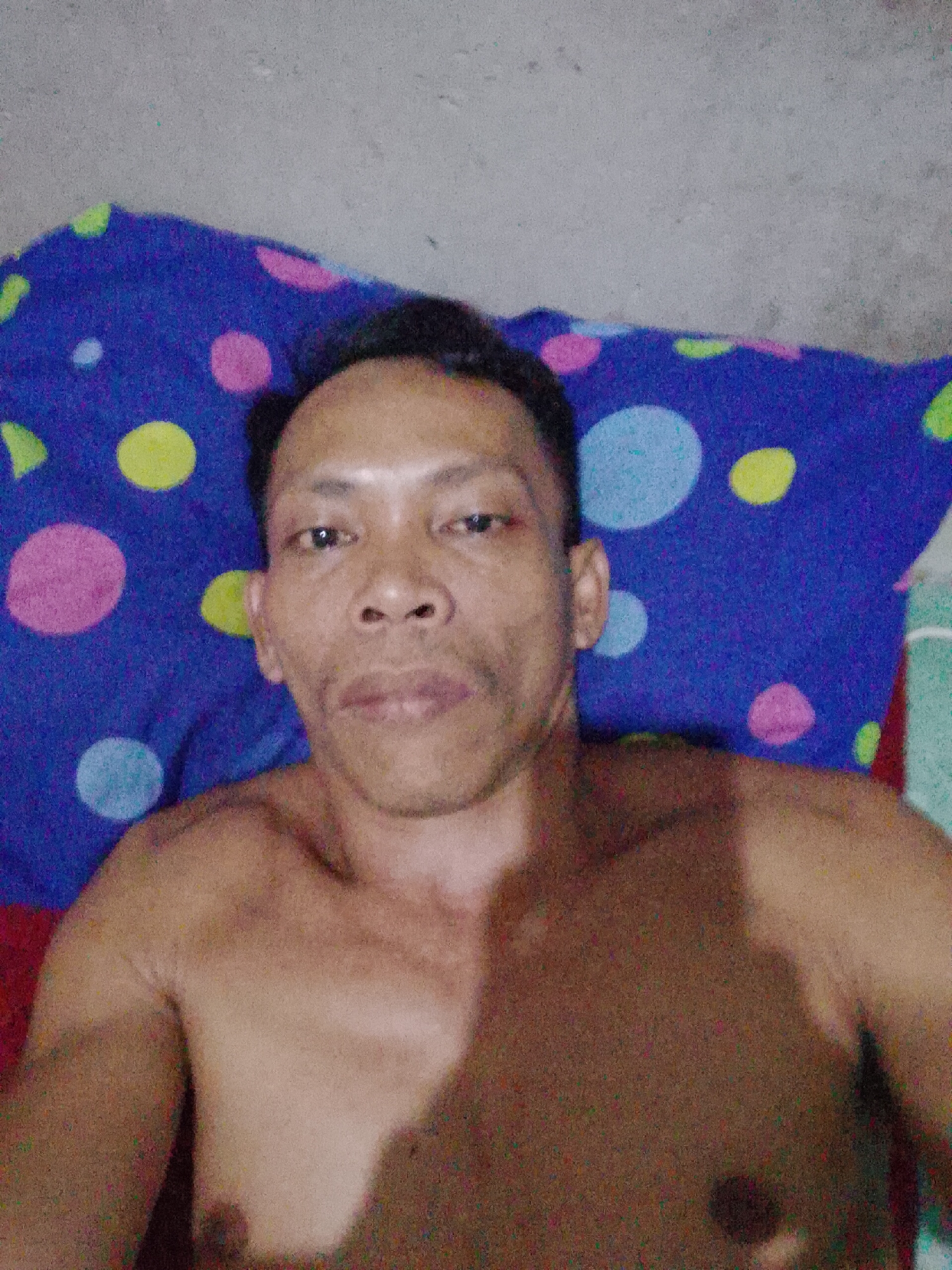 A shirtless man is lying on a bed with a blue polka dot pillow. He is facing the camera. The setting appears to be indoors, likely a bedroom, located in Bukit Ketok, Indonesia. The background is a plain grey wall.