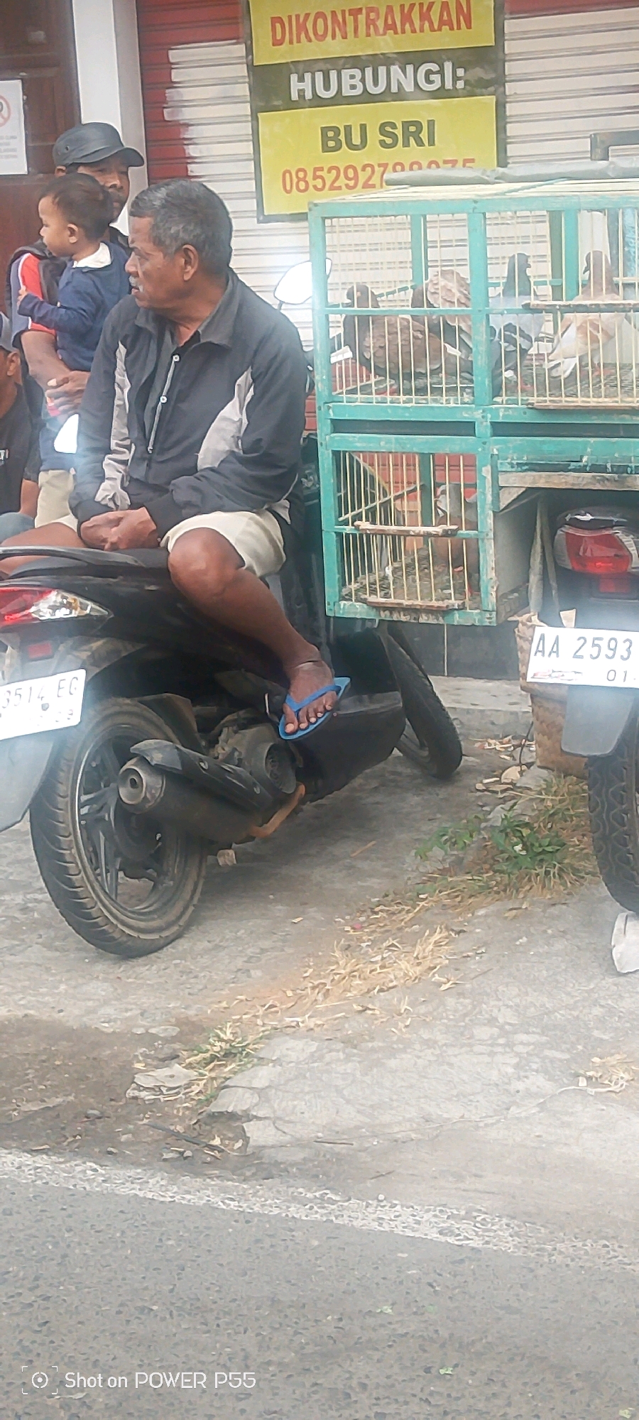 An outdoor medium shot captures a group of people and a birdcage mounted on a motorcycle. In the foreground, a man with graying hair and a dark jacket sits on a black motorcycle, wearing white shorts and blue flip-flops. His right leg is extended, with his foot resting on the ground. Behind him, a child in a blue shirt is held by another man wearing a dark cap and a black shirt. To the right of the motorcycle is a green birdcage filled with pigeons. A sign with Indonesian text hangs in the background, and a license plate reading "AA 2593 01" is visible on the back of the motorcycle. The ground is paved with a rough, gray surface, and dry vegetation is scattered around. The scene appears to be in a city setting.