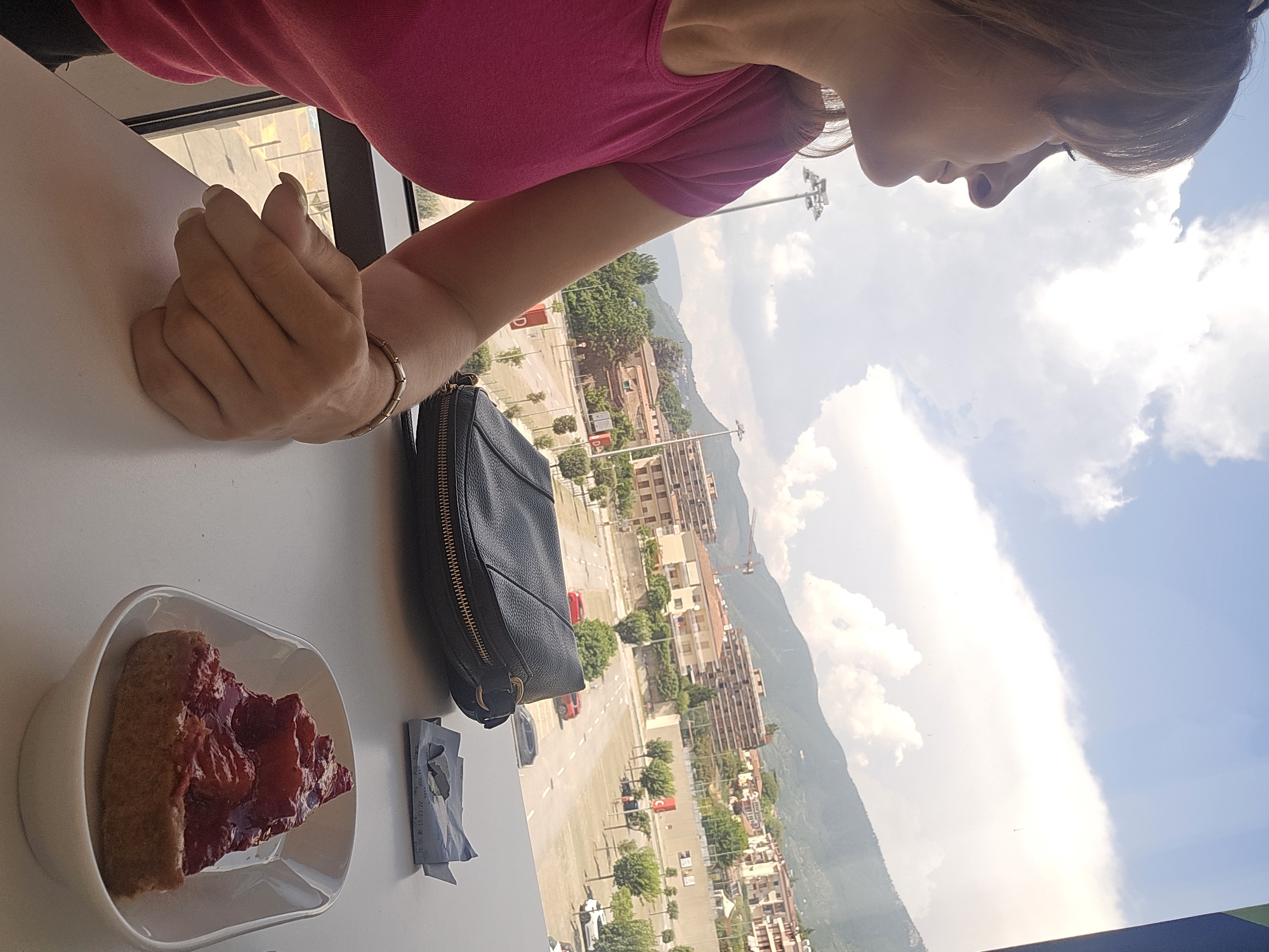 The image depicts a woman in profile, looking out a window. She has brown hair and is wearing a pink t-shirt. Her left hand is resting on a white table, and she is wearing a bracelet. To her right, on the table, is a slice of strawberry tart in a white bowl, a black purse, and a crumpled receipt or piece of paper. The window reveals a cityscape with buildings, trees, streetlights, and a hilly landscape in the background under a cloudy sky. The view suggests the location is in or near Baronissi, Italy, given the architectural style and the presence of the Italian flag visible on some of the buildings in the distance. The scene appears to be set indoors, likely a cafe or restaurant with a view.
