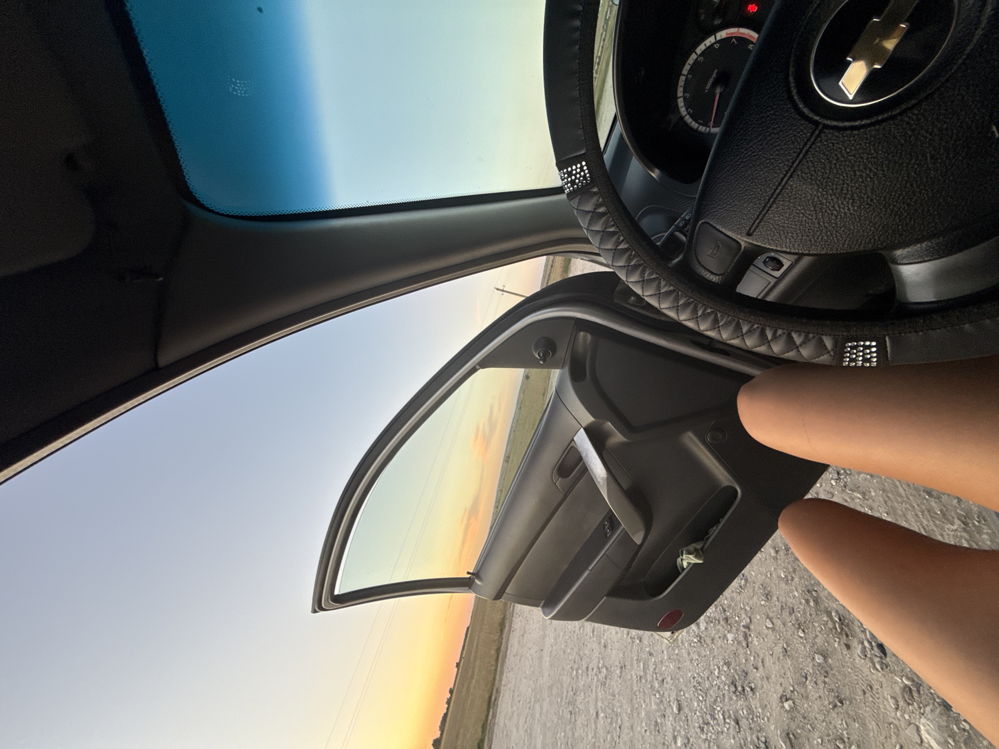 The image captures an interior view of a car, likely a Chevrolet based on the emblem on the steering wheel. The driver's side door is open, revealing a rural landscape with a dirt road and a field under a soft, pastel sky, suggesting either dawn or dusk. The sun's warm glow illuminates the horizon with hues of yellow and orange. Inside the car, the focus is on the steering wheel, which is covered with a black, quilted material and adorned with rhinestones. The dashboard is visible, including a speedometer. In the foreground, a person's bare legs are positioned towards the open door, implying they are either about to exit or have just entered the vehicle. The scene appears to be set in a quiet, open area, potentially on the outskirts of Cimișlia, Moldova, given the provided location context, characterized by its natural scenery and limited urban elements.