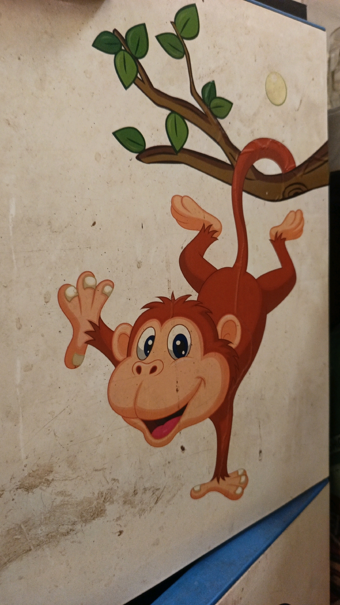 The image shows a cartoon illustration of a red monkey hanging from a tree branch. The monkey has a wide smile, big blue eyes, and is reaching out with one hand. The branch has green leaves. The illustration appears to be painted or drawn on a textured, light-colored surface, possibly a wall or a piece of furniture, with some visible dirt and scuff marks. A bright blue triangular shape is visible along the bottom edge of the image. The overall setting is unclear, and there is no information suggesting its location in North Kuta, Indonesia.