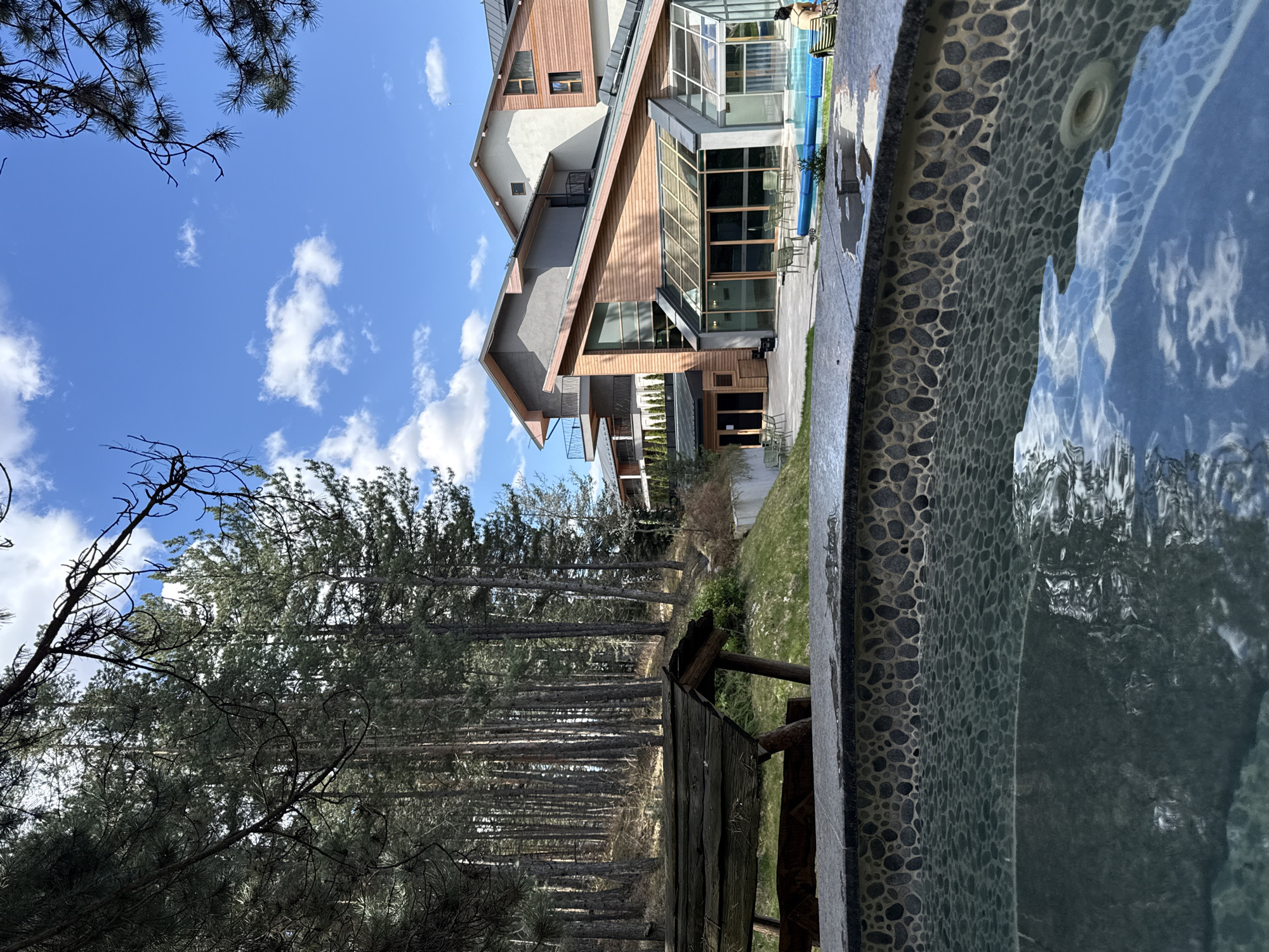 The scene depicts the external environment of a modern building, likely a hotel or resort, in Velingrad, Bulgaria. The building has wood and concrete paneling and a glass-enclosed section with chairs visible inside. To the left, there is a pine forest with tall, slender trees. In the foreground is a feature that appears to be the edge of a pool or hot tub, with a patterned exterior. The sky is visible above, showing a clear blue sky with some clouds. There are no people visible in the scene. A wooden structure, possibly a gazebo or small shed, is partially visible near the forest. The image gives a sense of the resort being nestled within a natural environment.