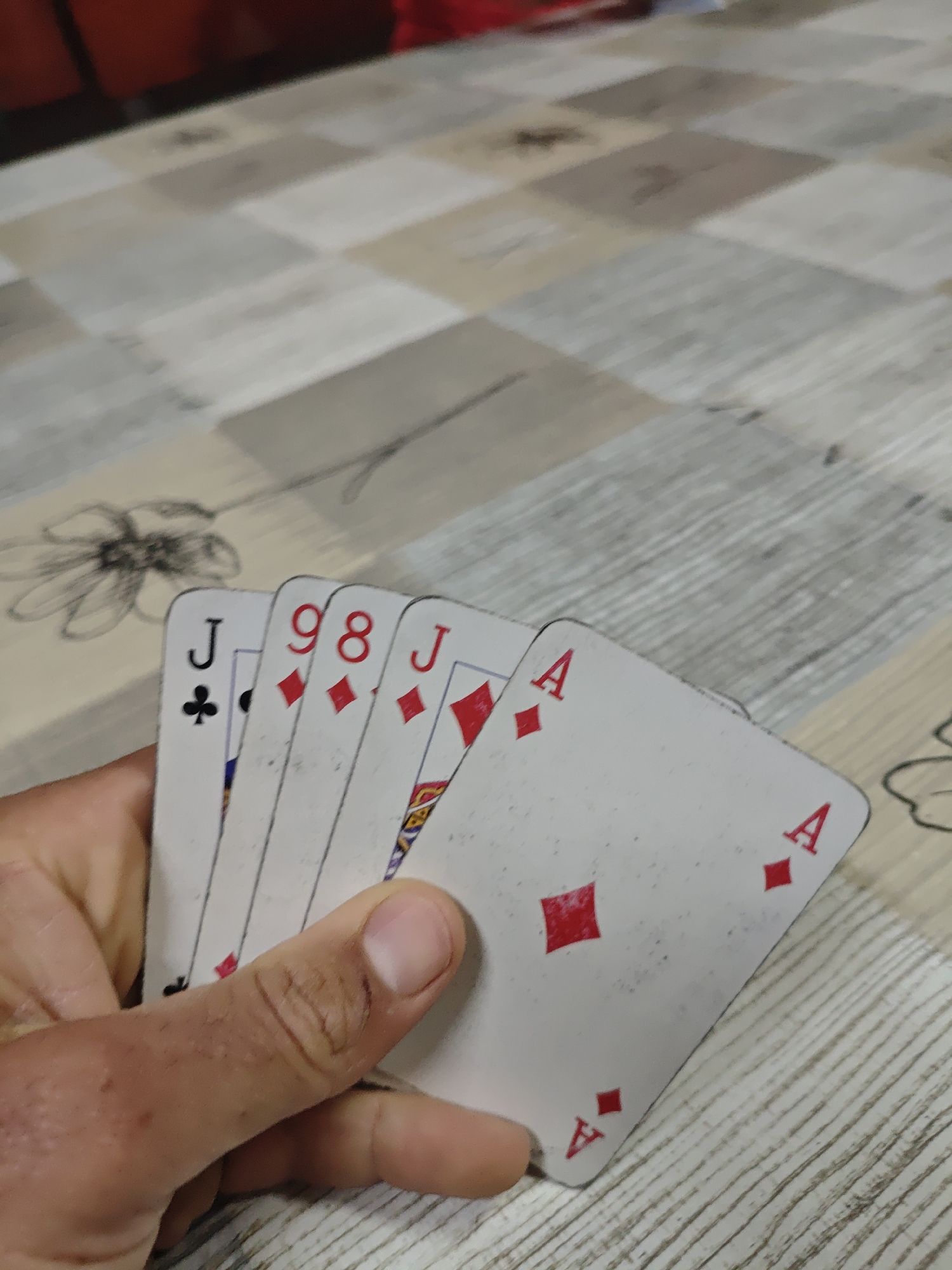 A person is holding five playing cards - a Jack of Clubs, an 8 of Diamonds, a 9 of Diamonds, a Jack of Diamonds, and an Ace of Diamonds. The cards are being held over a table with a patterned surface, featuring a floral design. The location is Unknown, Unknown.