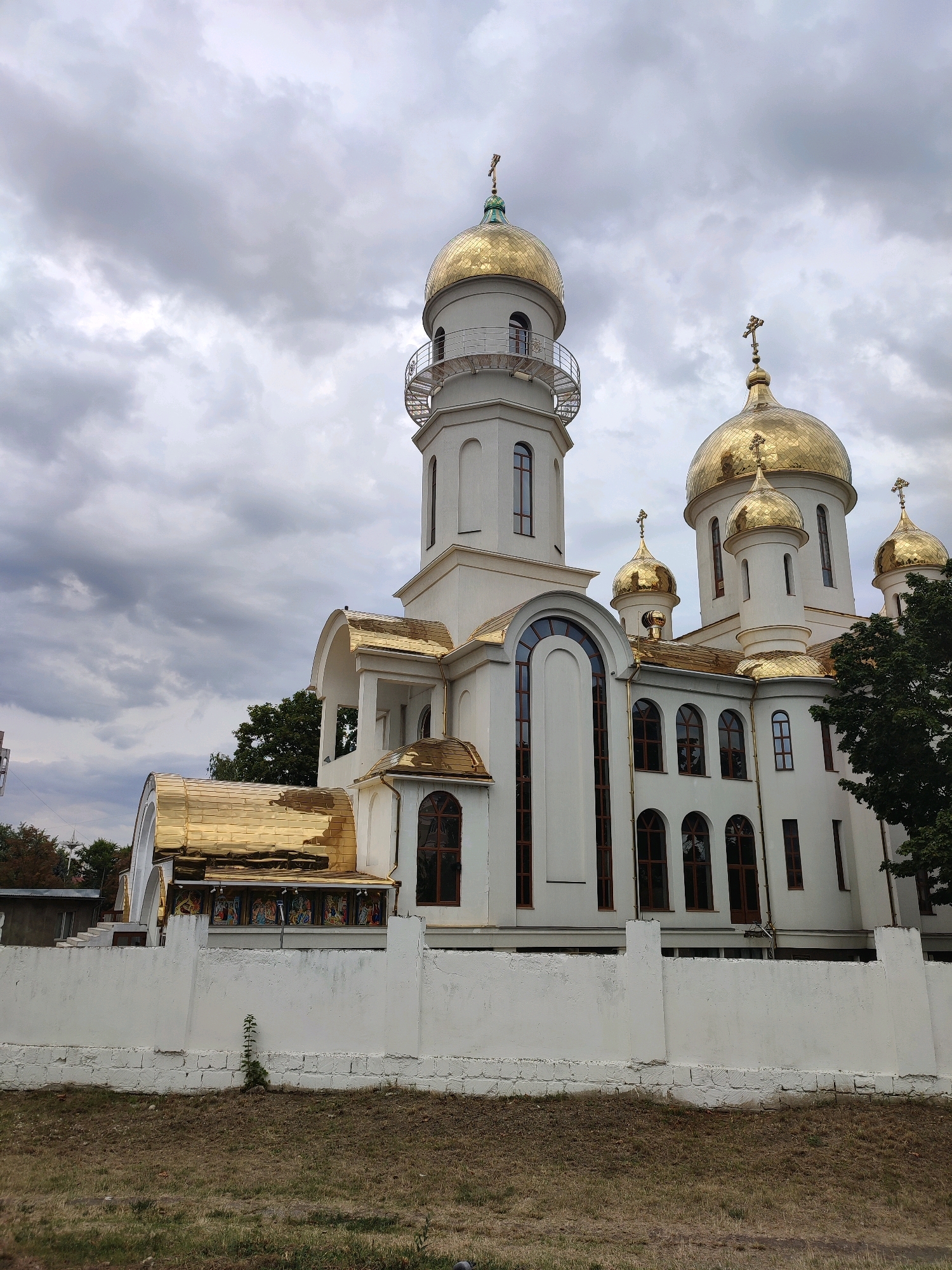 The image shows an exterior view of a white Orthodox church with golden domes and crosses. The church has several distinct sections, including a tall, narrow tower with a balcony and a larger main building with multiple arched windows. A white stone wall runs along the front of the property, with dry grass and some sparse vegetation in the foreground. The sky is overcast with heavy clouds. The church is identified as the Nativity Cathedral in Chișinău, Moldova.