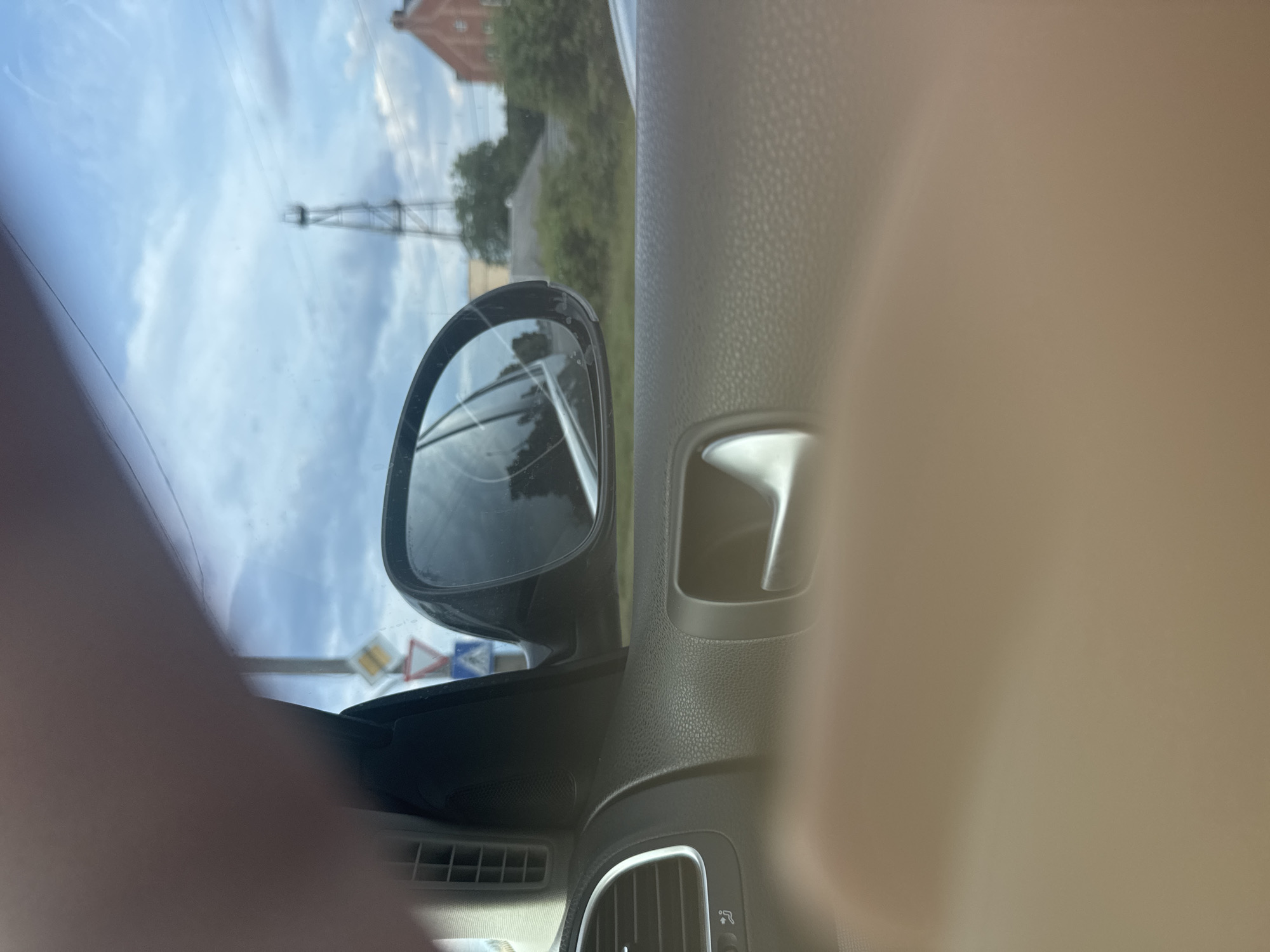 The image depicts a view from the interior of a vehicle, looking outwards through the passenger side window and reflecting in the side mirror. The foreground on the bottom and top-left is out of focus, showing blurred, skin-toned areas, likely a hand or arm obstructing the view.
Inside the vehicle, visible details include a segment of the dark grey, textured dashboard and door panel. On the far left, a vertical air vent is present, and below it, a portion of a control dial featuring a fan symbol and "AC" lettering is discernible. Towards the center-right, a silver-colored interior door handle is visible. The exterior side mirror, positioned on the right, is black and shows multiple distinct cracks across its reflective surface.
Outside the vehicle, through the window, the scene is somewhat blurry due to the glass and distance. On the left, a set of road signs is mounted on a pole: a white sign with a yellow diamond (indicating a priority road), directly below it a red triangle sign pointing downwards (suggesting 'yield'), and a partially obscured blue sign further down. Beyond the mirror, a tall, slender metal lattice structure, possibly a utility pylon or communication tower, stands against an overcast sky. Further to the right, a reddish-brown brick building with a pitched roof is partially visible, nestled amongst green foliage. The sky is largely covered with clouds. In the far bottom-right, a sliver of what appears to be a light-colored vehicle is discernible. The overall environment suggests an urban or semi-urban setting in the city of Unknown, Unknown, under daylight conditions.