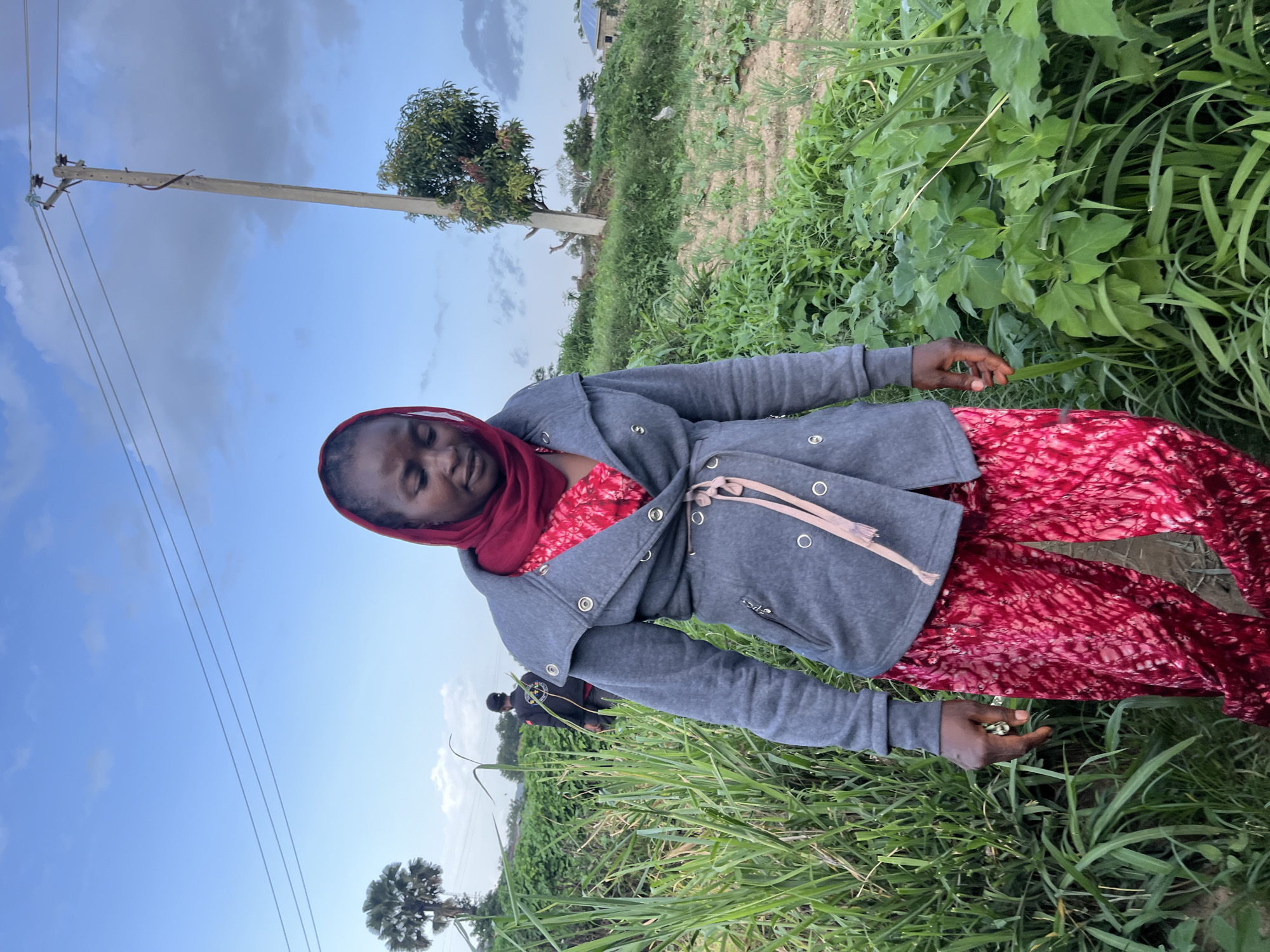 A young Black woman wearing a red headscarf and a grey jacket over a red dress stands in a field of tall green grass under a blue sky with scattered clouds. Her eyes are closed, and she appears to be relaxing or in thought. Her right hand is extended, and she is gently holding a few green pods, possibly from a plant in the field. The setting appears to be rural, with a utility pole and wires visible in the background, suggesting a residential or agricultural area. The sun is out, indicating it is likely daytime. The overall mood is peaceful and serene.
