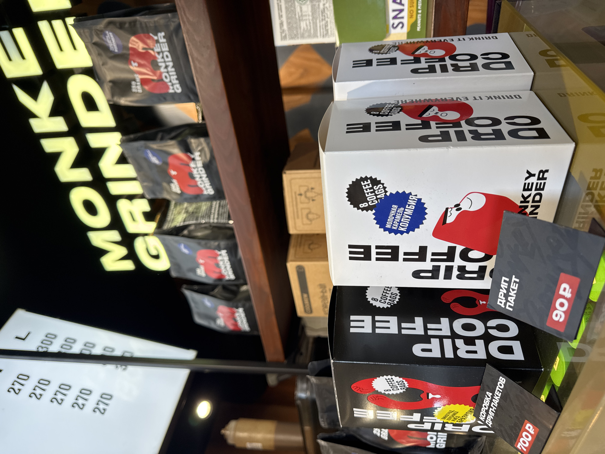 The image shows a display shelf in a store, likely located in Perm, Russia, as evidenced by the Russian text on the product labels and price tags. The shelf primarily features "Monkey Grinder" brand coffee products, including 250g bags of beans and "Drip Coffee" boxes containing 8 coffee bags each. There are two varieties of the drip coffee: "Brazil Grape" and "Milk Caramel Columbia." The price tags indicate that a box of drip coffee costs 700 rubles and a single drip packet is 90 rubles. The "Monkey Grinder" branding is also prominently displayed on a sign. The shelf is made of dark wood, and the products are arranged in an organized fashion. There's a price list with numbers on a white label and some food snacks above.