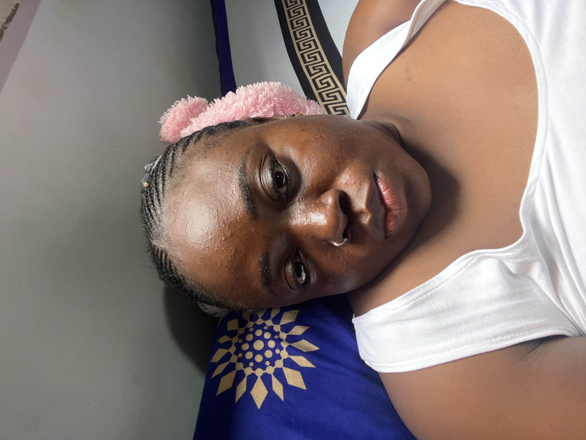 A close-up, head-and-shoulders selfie of a Black woman with braided hair and a pink accessory on her head is pictured. She is wearing a white tank top and has a silver nose ring. The background features a pillow with a blue and gold sun-like design and another pillow with a black and gold geometric pattern. The scene appears to be indoors, possibly in a bedroom. The lighting is somewhat dim, casting shadows on the woman's face. The overall impression is casual and intimate, suggesting a personal moment captured by the subject herself. The image does not provide any specific visual cues to place it within the city of Abuja, Nigeria.