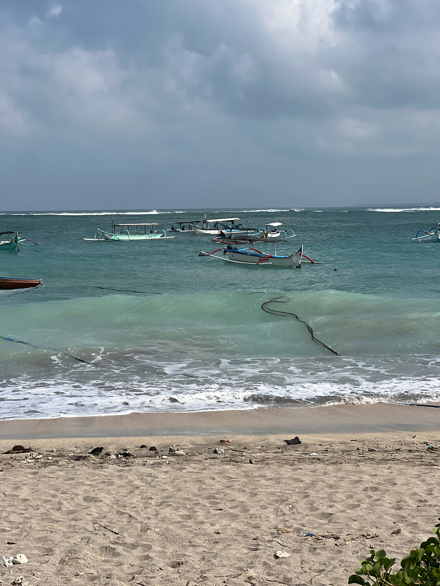 A medium shot captures a serene beach scene in Kuta, Indonesia, under a cloudy sky. Several traditional Balinese boats, known as jukung, are moored in the turquoise waters, their outriggers visible against the gentle waves. One boat, painted white with blue trim and red accents, sits closer to the shore. A dark rope snakes through the water, connecting to a boat or anchor. The foreground features a sandy beach littered with various debris, including shells, driftwood, and plastic waste. Lush green foliage is visible on the right side of the frame. The ocean stretches to the horizon, where white-capped waves break, creating a sense of depth and vastness. The overall atmosphere is peaceful, with the natural beauty of the beach contrasting with the signs of human activity and environmental impact.