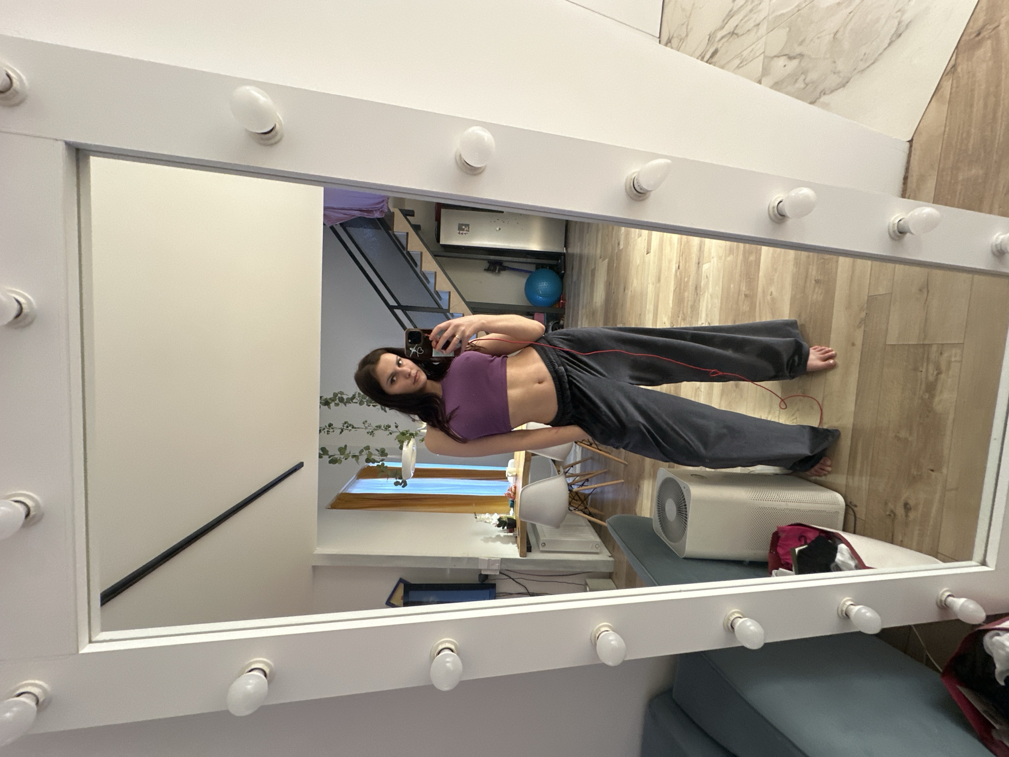 A young woman with long dark hair and a light complexion is taking a selfie in a large, brightly lit mirror. She is wearing a purple crop top and dark gray sweatpants. Her pose is casual, with one hand holding her phone and the other resting on her hip. The mirror is framed by a white border with numerous light bulbs, suggesting it's a vanity mirror.
The reflection shows a room with light-colored walls and wooden flooring. In the background, there's a staircase leading up, a blue exercise ball, and a white air purifier. There are also some plants on a shelf. The overall impression is of an indoor space, possibly a dressing room or a home gym. The lighting from the mirror suggests it could be daytime, or the lights are on for ambiance or use. The woman's expression is neutral and direct, looking at her reflection as she takes the photo. There are no other people visible. The image is oriented vertically, with the top of the mirror frame appearing at the bottom of the frame.