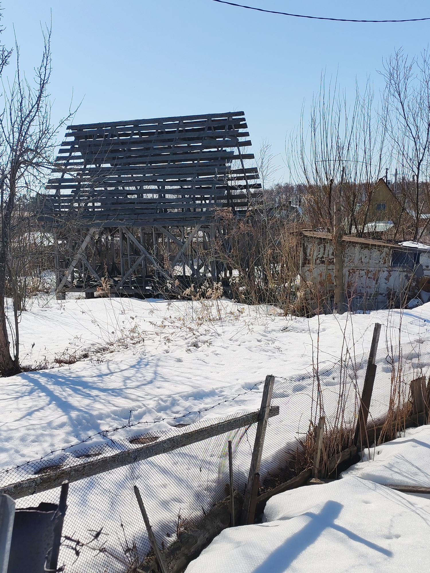 The image depicts a snow-covered scene in an area likely in or near Chaikovsky, Russia. A partially constructed or dilapidated wooden structure dominates the midground. The structure consists of a skeletal frame, likely of a building, with exposed beams forming its roof and walls.
In the foreground, a dilapidated wire fence runs across the frame, partially obscured by snow drifts. The area behind the fence is also covered in snow, with dried vegetation and a few bare trees scattered across the landscape. The scene is lit by bright sunlight, casting long shadows across the snow. A rusty, damaged trailer or shed is visible behind the wooden structure, further indicating a state of disrepair and abandonment. In the background, a few houses or buildings can be seen, hinting at a residential area. The sky is clear and blue. No people are visible in the image.