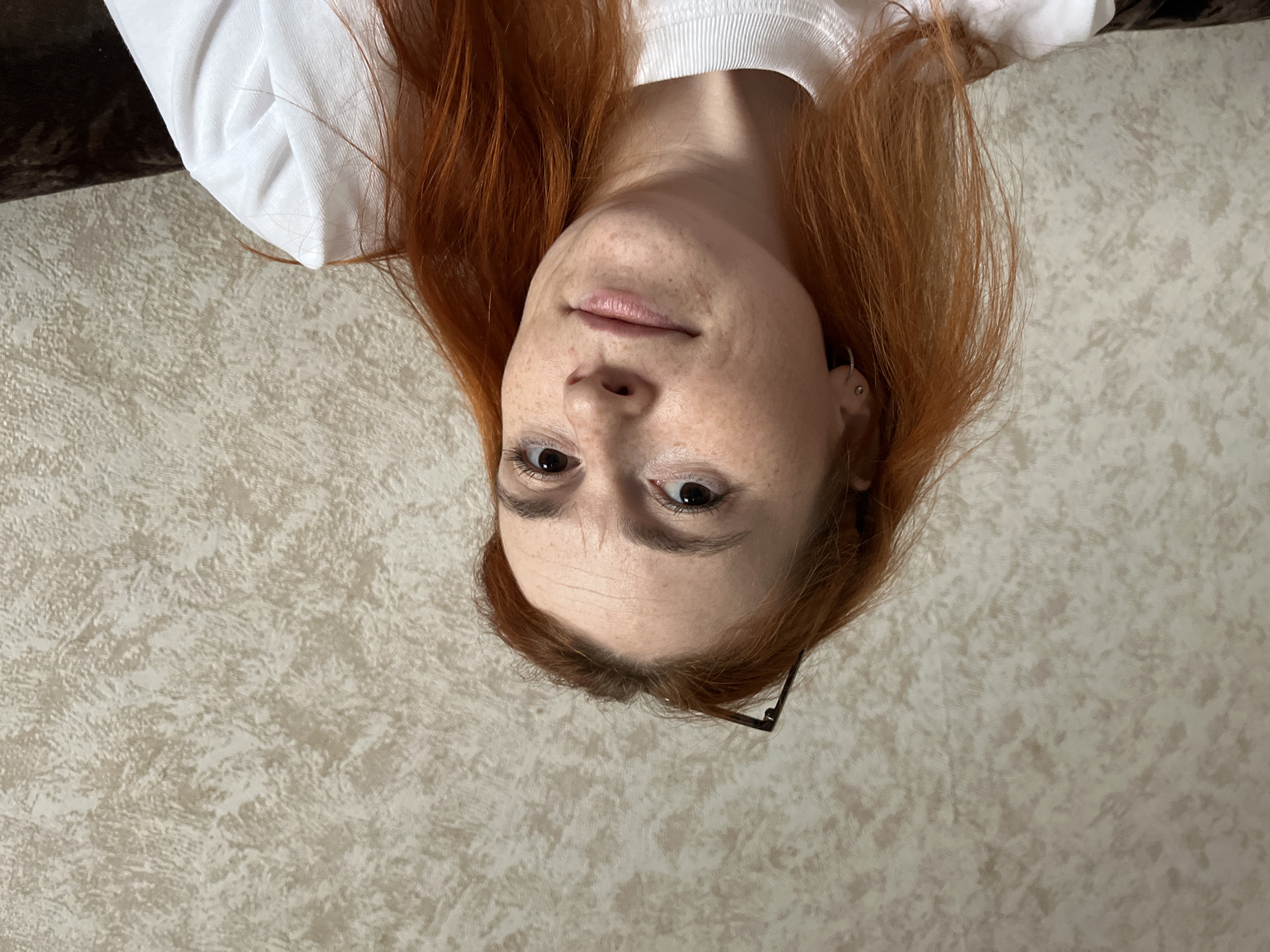 The image is a portrait of a young woman with red hair and freckles. She is lying on her back, and the camera is angled from above, creating an upside-down perspective. She is wearing a white t-shirt. Her eyes are open and looking towards the camera. The background is a textured wallpaper. The lighting appears to be from an indoor source. There is no visible text in the image.