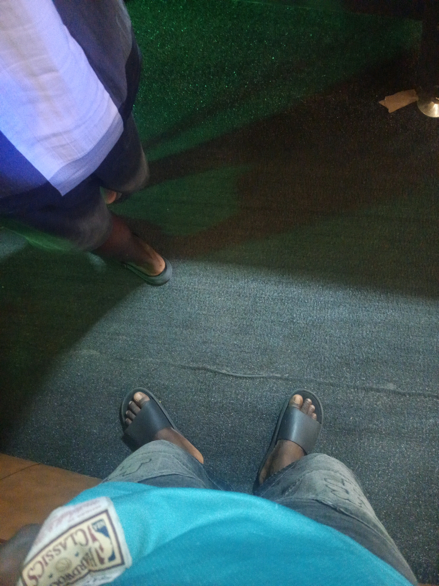 The image shows a person's legs and feet, as well as a portion of their torso, from a first-person perspective. The person is wearing black slide sandals and appears to be standing on a textured, dark-colored surface. In the background, there is a patch of green, possibly artificial turf or a carpeted area, with a shadow cast upon it. To the left, a portion of what appears to be clothing, with stripes of white and dark fabric, is visible. The overall setting suggests an indoor or outdoor location with artificial lighting, possibly a recreational area or a commercial establishment. The scene is static, with no apparent activity beyond the person's presence. This image does not provide specific visual cues to definitively place it within the city of Obinze, Nigeria.